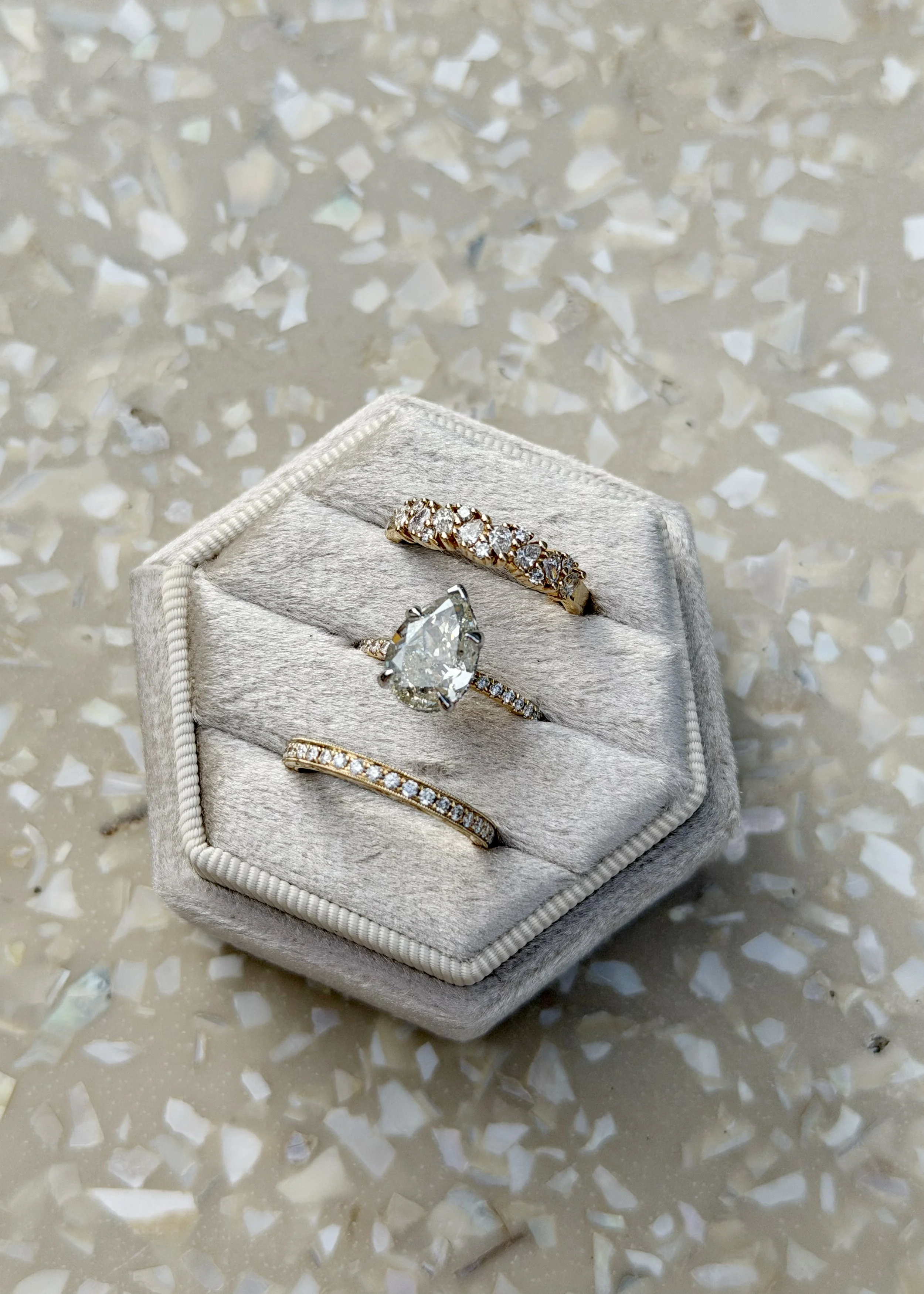 Three 14k yellow gold rings in a velvet ring holder box. The three rings show distinctly unique stone settings, with the middle ring being a diamond engagement ring with a pear cut center stone and diamond pave around the band.