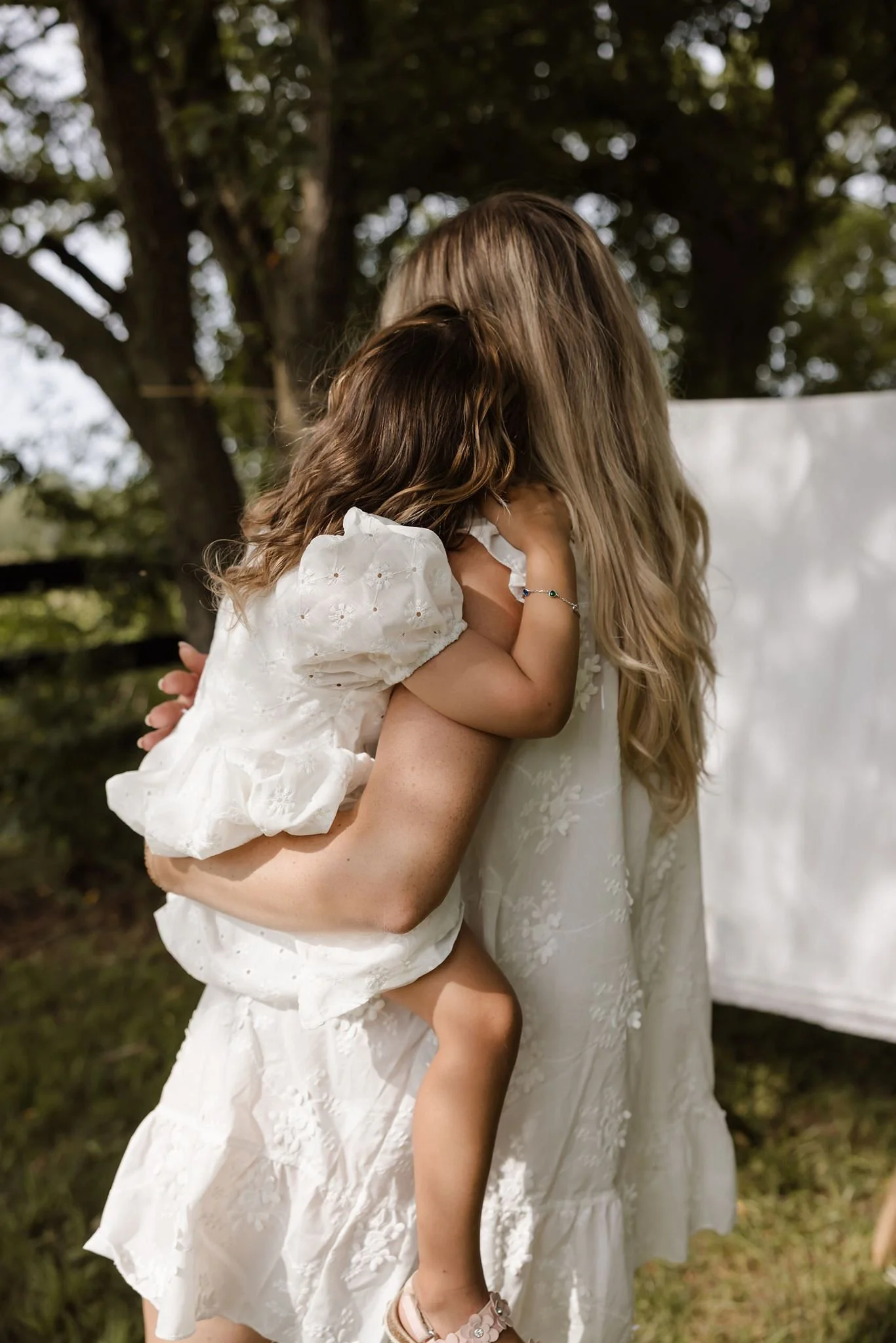 A mother carrying her young daughter with their backs turned to the camera as they gaze upon the garden. The young girl is wearing a sterling silver bracelet with sapphire gemstone stations from Brilliant Fine+Custom Jewelry