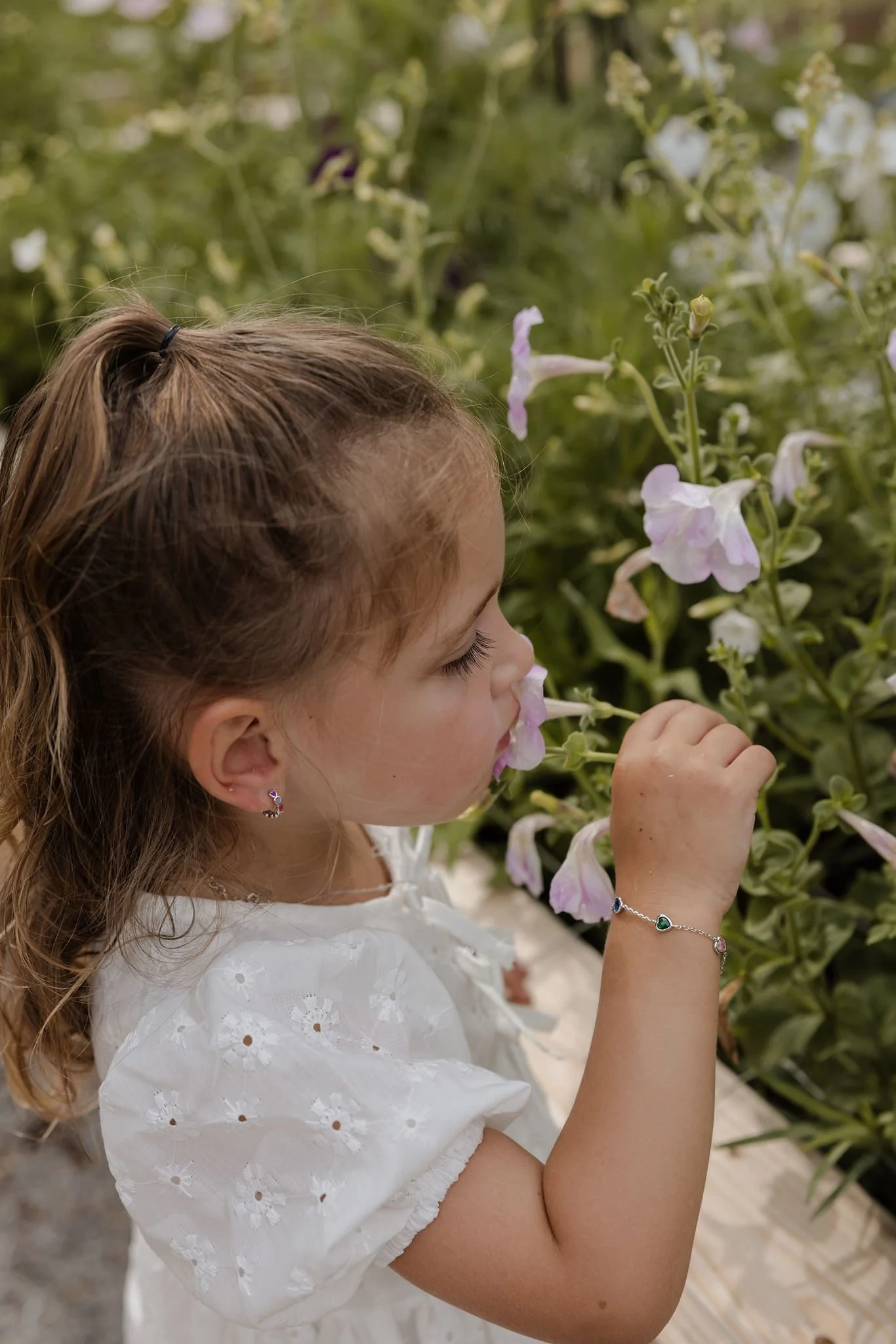 A young girl smells a flower in a garden, she is wearing small enamel heart huggy earrings from Brilliant Fine+Custom Jewelry