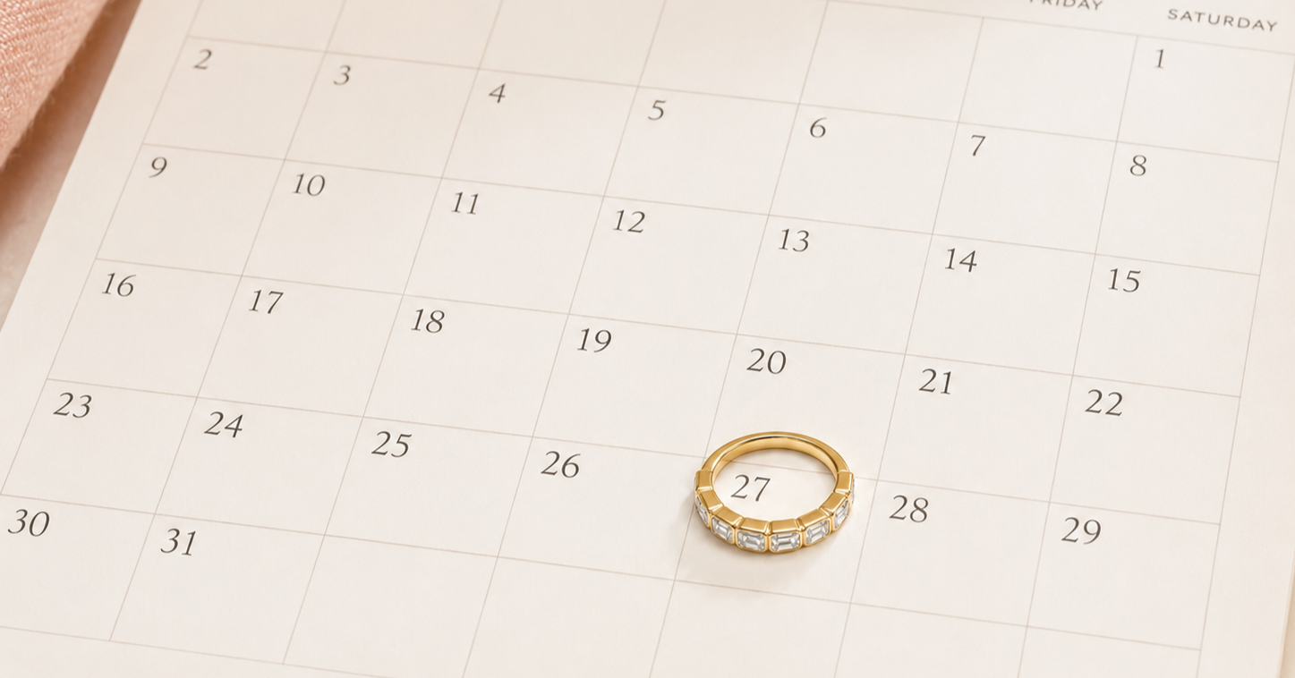 A gold wedding band with diamonds set in half the band sitting on a calendar encircling the date of August 27th - The date of our "Stack Your Story" special event at Brilliant Fine+Custom Jewelry