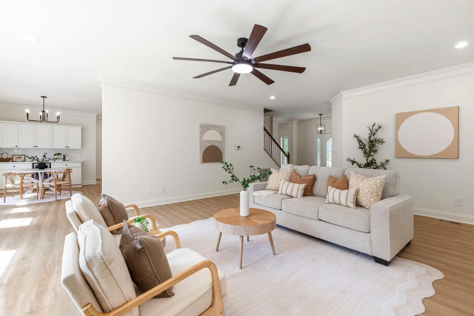Living room with white walls, light wood flooring, beige sofa with pillows, wooden coffee table with a white vase and greenery, wall art, and a large ceiling fan. Open view to kitchen and staircase.