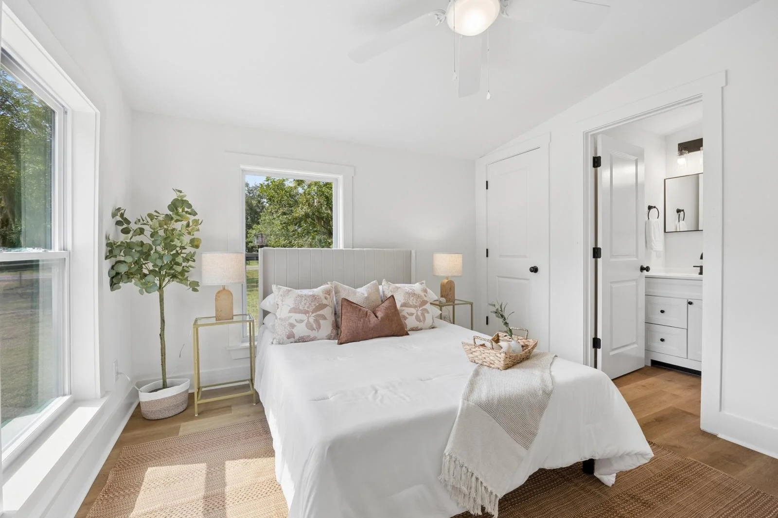 Bright bedroom with white walls, large window, bed with white bedding and decorative pillows, two bedside tables with lamps, a potted plant, and a view of trees outside.