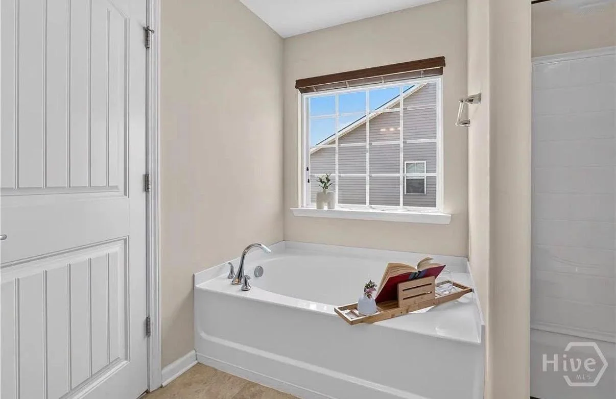 A bathroom with a white bathtub next to a window with blinds. There is a wooden tray on the tub holding an open book, a small vase with flowers, and some glasses. A potted plant is on the window sill, and a part of a shower enclosure is visible on th