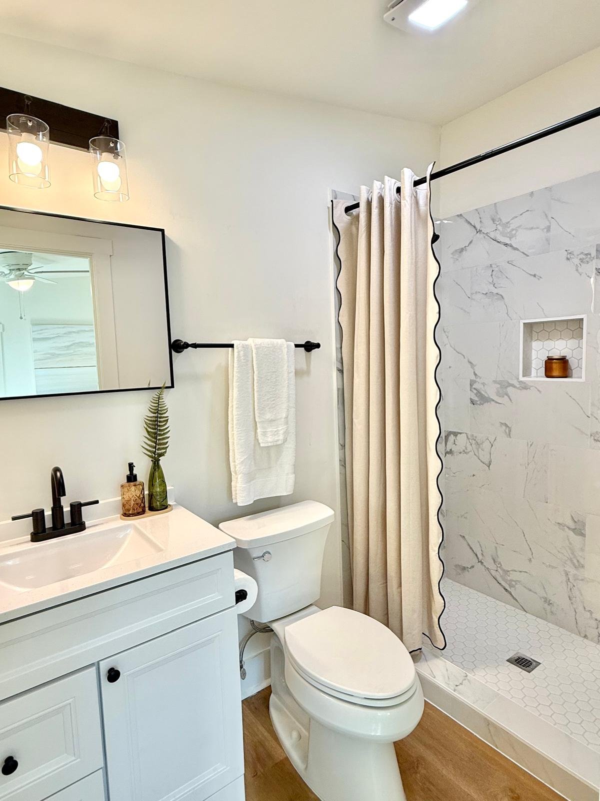 A modern small bathroom with a white vanity, black faucet, large mirror, framed light fixtures, a white towel on a black towel bar, a toilet, a shower with a beige curtain, marble tile walls, a small built-in shelf with a brown jar, and wood flooring