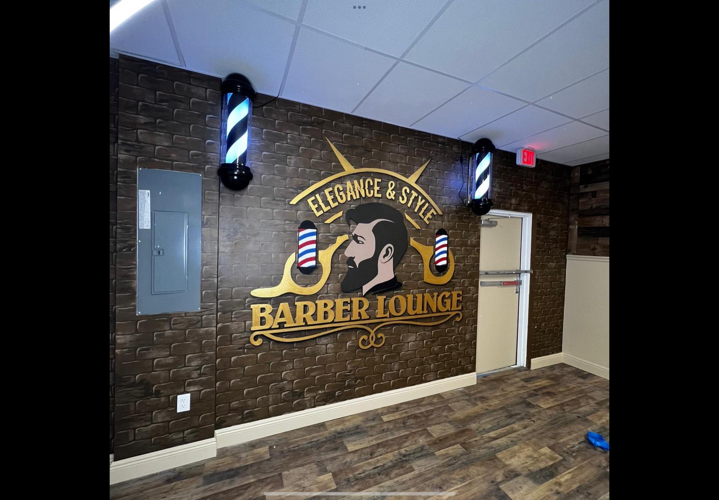Interior view of a barber lounge with a rough brick wall, a salon logo featuring a man's profile with styled hair and beard, surrounded by barber tools, and lighting fixtures with barber pole patterns.