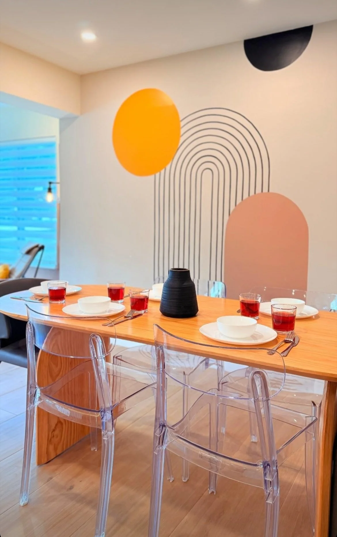 A dining table set with four white bowls, four white plates, four forks, and four glasses filled with a red liquid. A black vase is placed in the center of the table. The chairs around the table are transparent. In the background, there is a wall with modern geometric art featuring large orange, pink, and black shapes with black lines arching over the shapes.