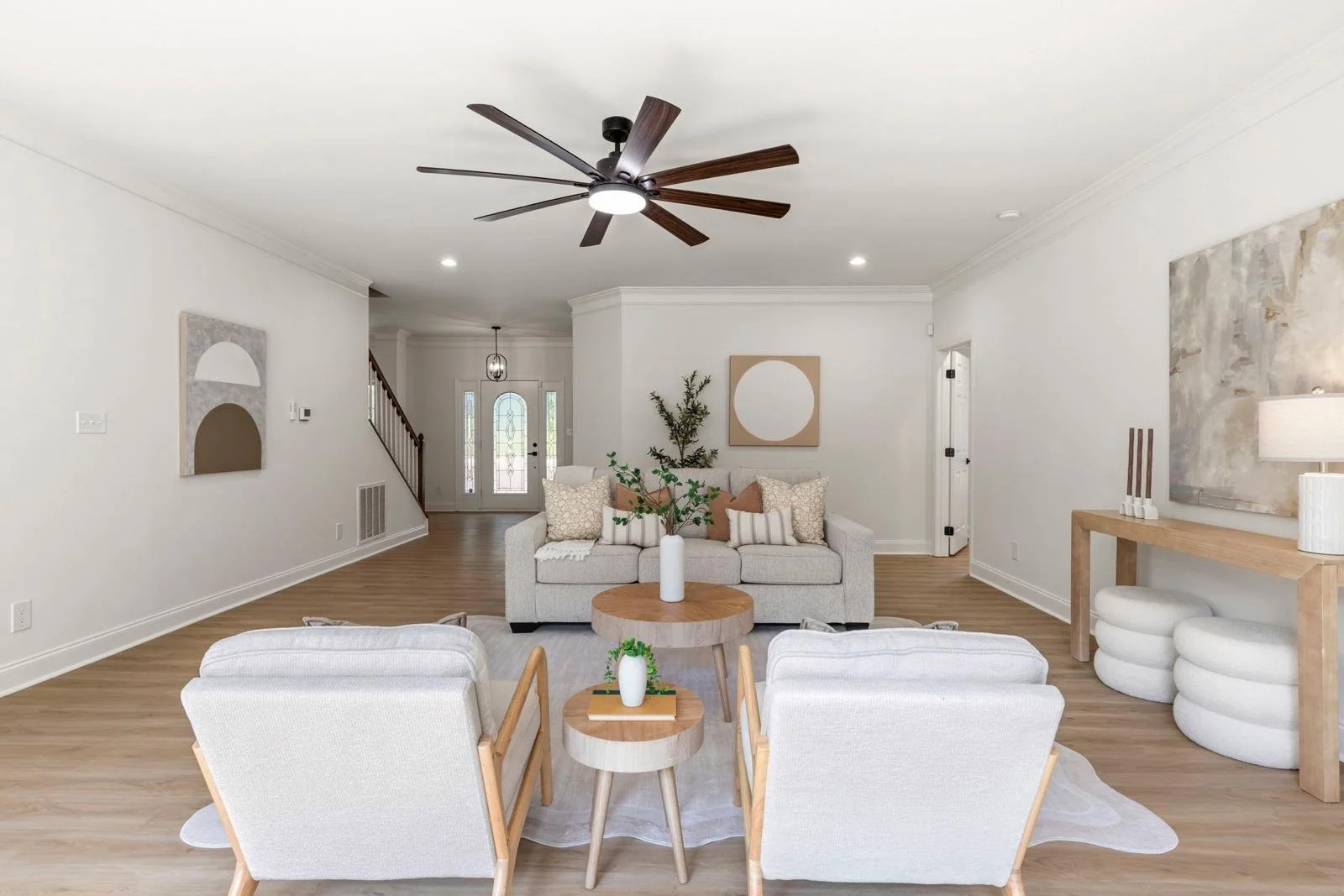 Interior of a modern living room with light-colored walls, hardwood floors, a ceiling fan, and minimalistic furniture including a sofa, armchairs, and side tables decorated with vases and plants, with artwork on the walls and a front door visible in 