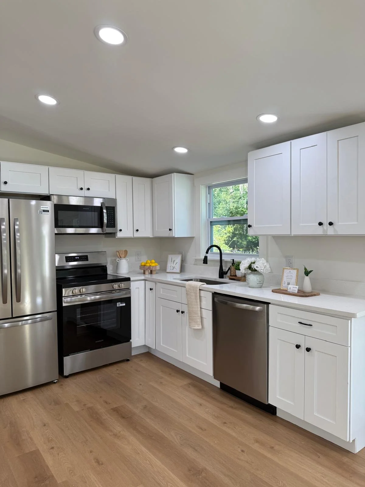 A modern kitchen with white cabinets, stainless steel appliances including a refrigerator, microwave, oven, and dishwasher. There is a black sink with a black faucet, a window with a view of greenery, and decorative items like flowers, framed picture