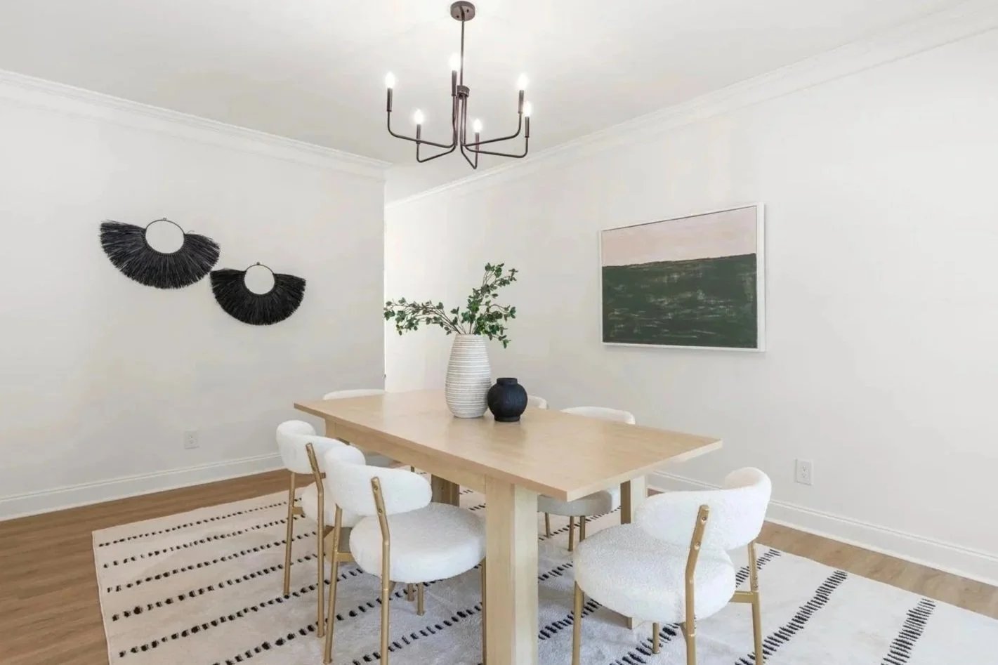 A modern dining room with a wooden table, six white upholstered chairs with gold accents, and minimalist decor including black and white wall art, a large vase with green branches, and a landscape painting on the wall. A black chandelier hangs from the ceiling over the table.