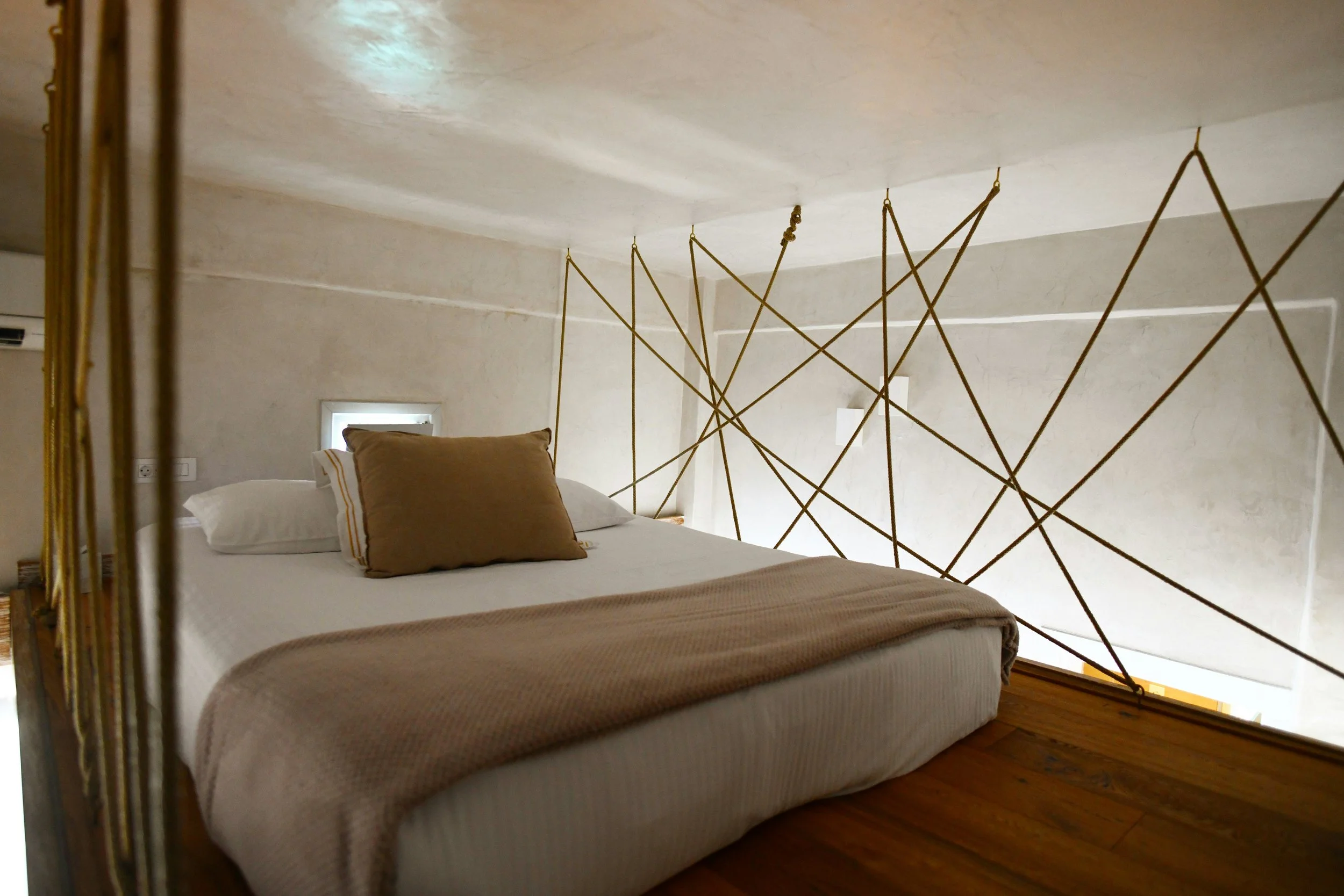 A minimalist bedroom featuring a white bed with beige blankets and pillows, a ceiling with textured white paint, and a decorative gold rope partition with geometric patterns dividing the space.
