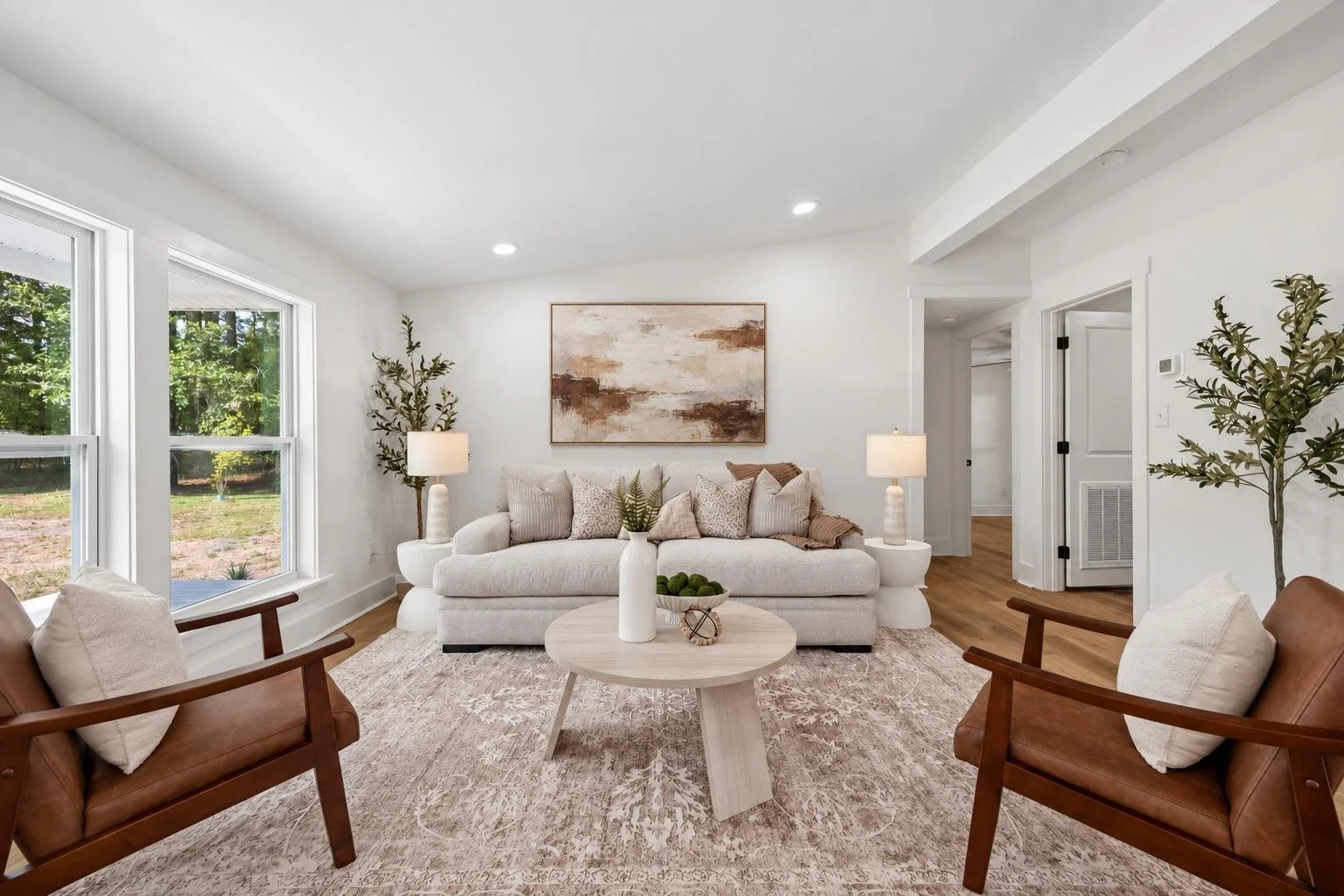 A living room with white walls, hardwood floors, a white sofa with pillows, two brown armchairs, side tables with lamps, a round coffee table with a vase and decorative objects, large windows showing outdoors, a framed artwork on the wall, and indoor