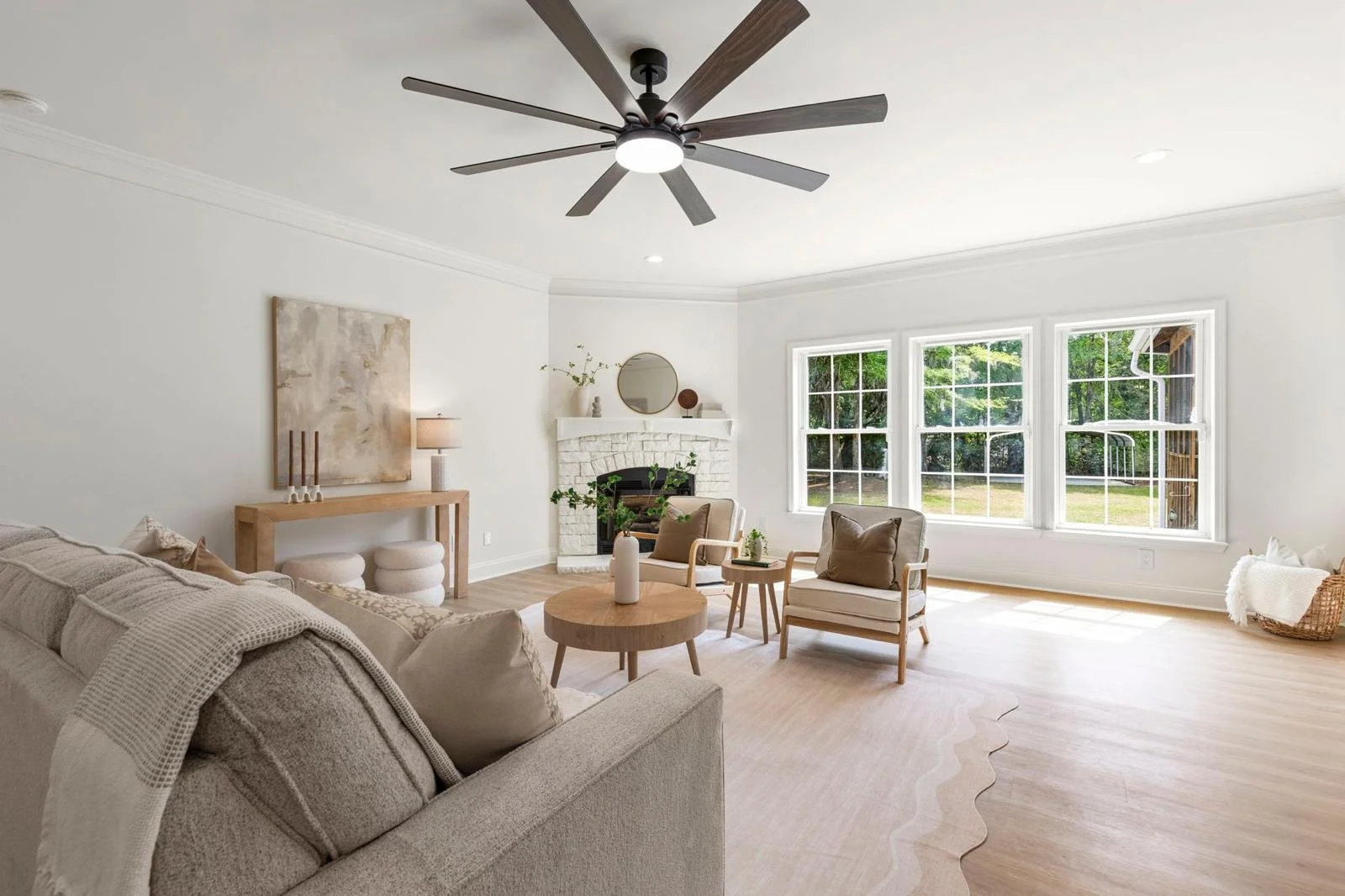 Bright living room with large windows, white walls, a neutral-toned sofa, wooden chairs, a round coffee table, a fireplace with a mirror and decorative items, and a ceiling fan with dark blades.