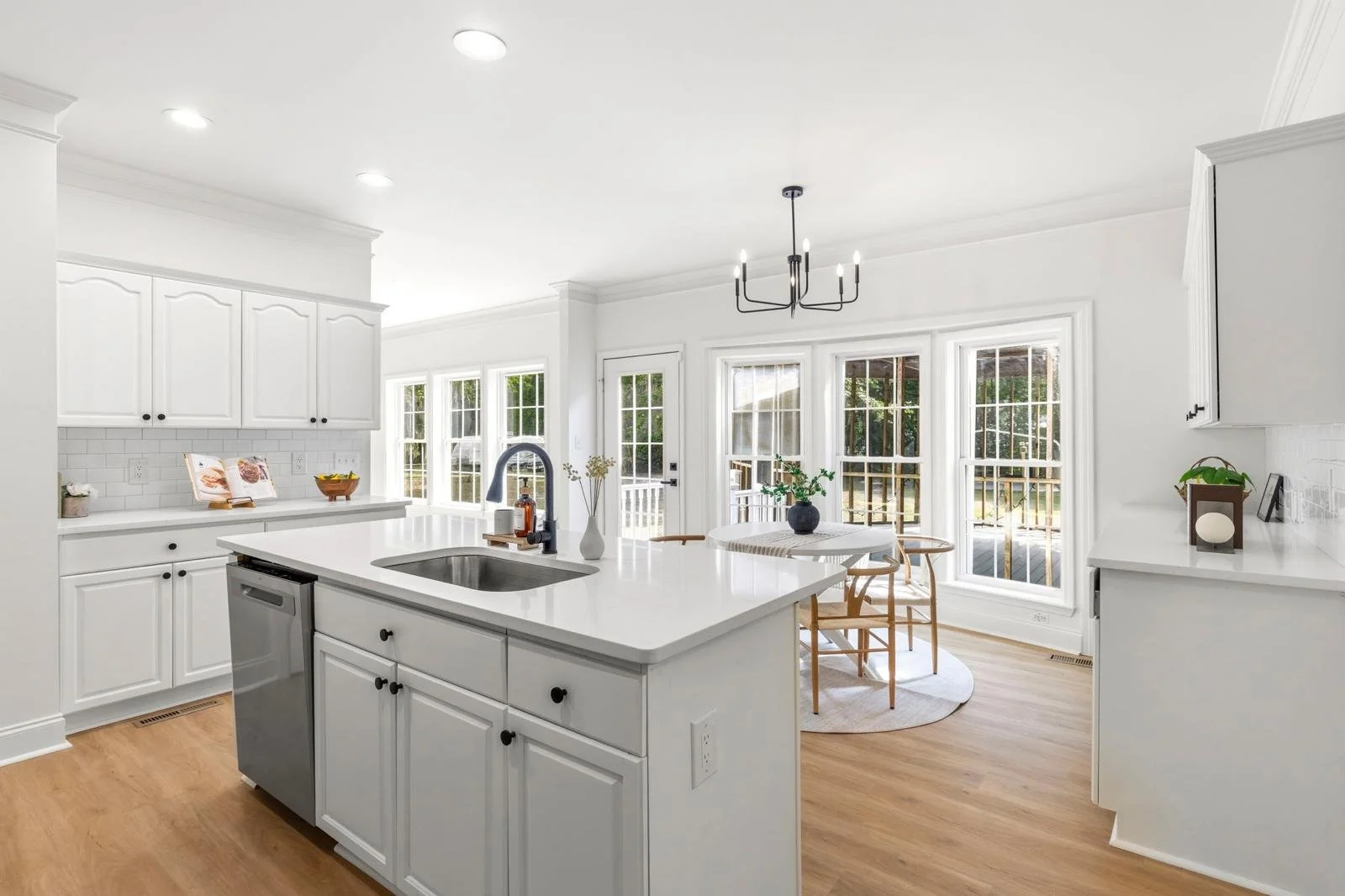 Bright, modern kitchen with white cabinets, island with sink, wooden floors, round dining table with chairs, large windows with natural light, black chandelier, and decor plants.