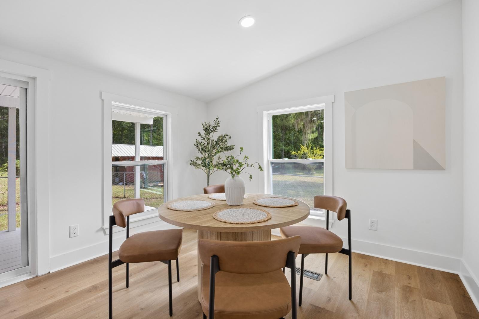 A dining area with a round wooden table, four beige chairs, a white vase with greenery, large windows showing an outdoor view, and a beige abstract artwork on the wall.