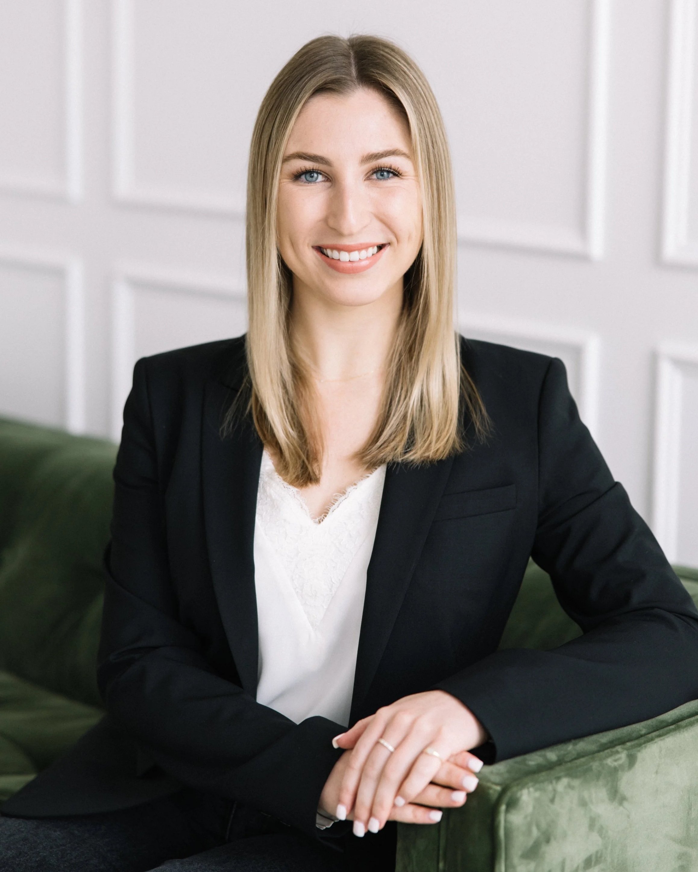 A young woman with blonde hair, blue eyes, and fair skin smiling while sitting on a green velvet sofa. She is wearing a light gray blazer and white top, with her left arm resting on the sofa's armrest. The background features white paneled walls.