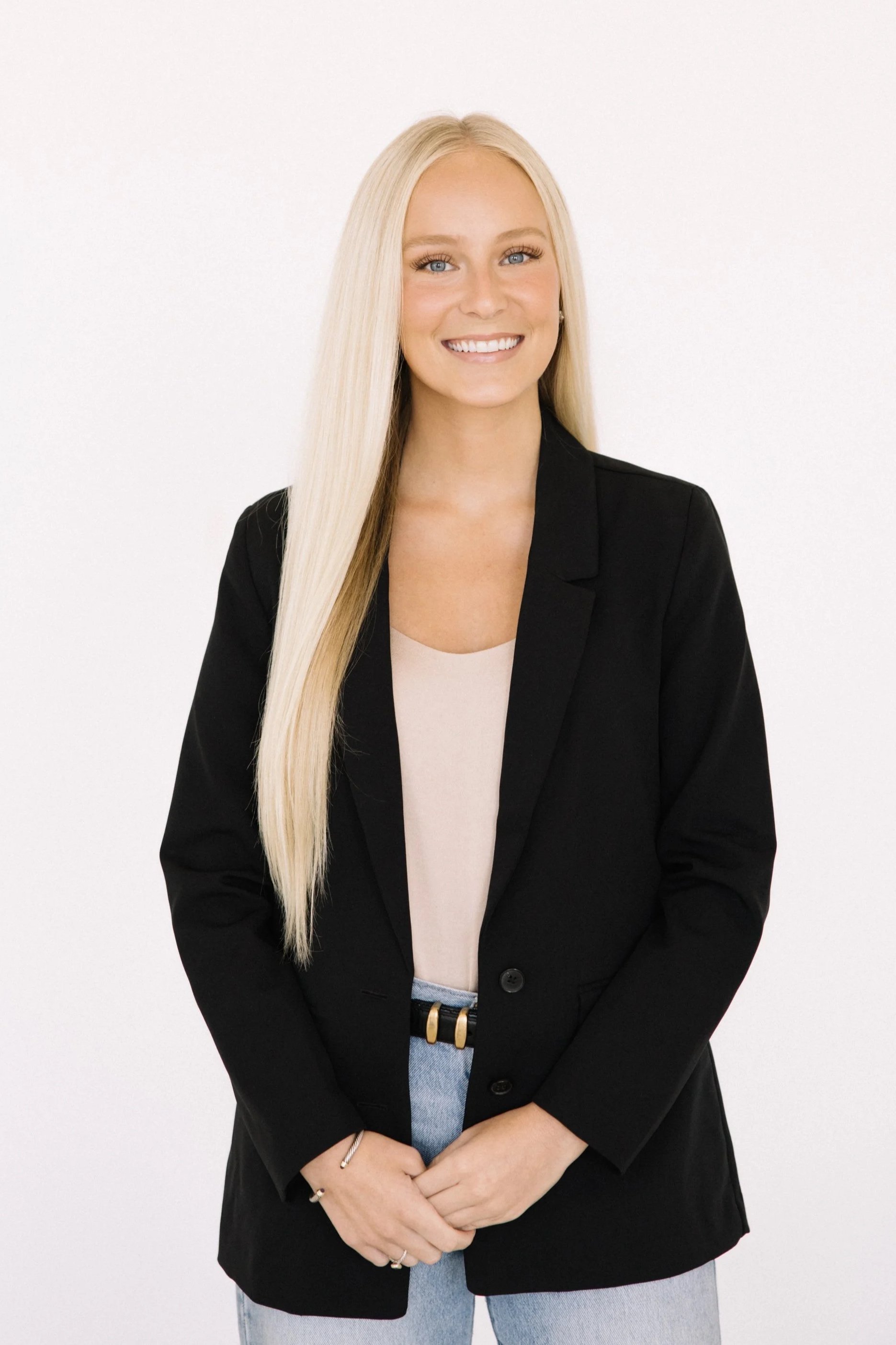 A woman with long blonde hair, blue eyes, and fair skin sitting on a green velvet sofa, smiling at the camera. She is wearing a black blazer, gold earrings, a gold watch, and a bracelet.