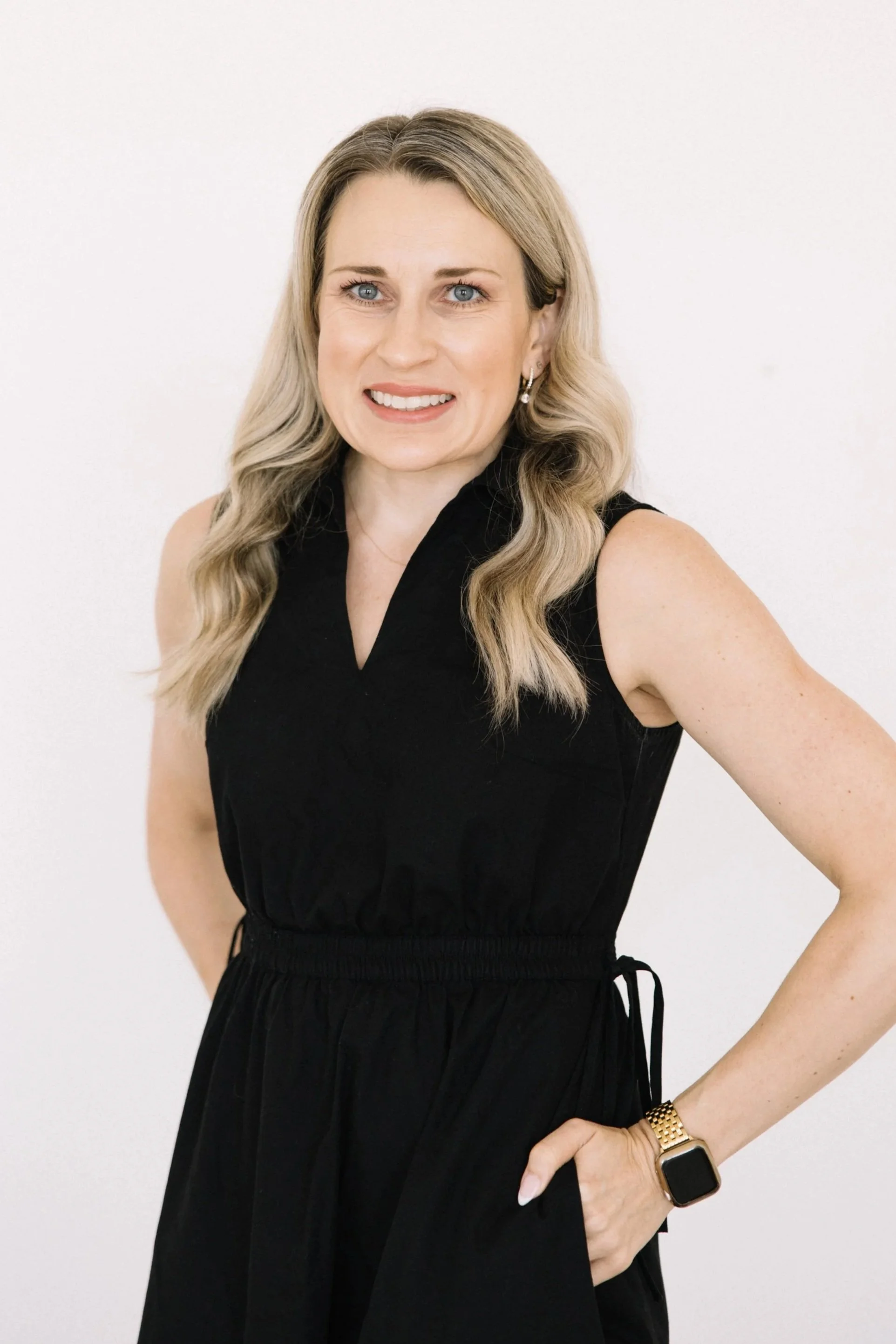 A woman with long blonde hair, wearing a black sleevless dress, standing, looking at the camera with a smile, with a white wall in the background.