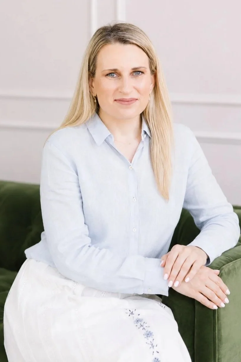 A woman with long blonde hair, wearing a blue blouse and white skirt with flowers, sitting on a green sofa, looking at the camera with a slight smile, with a white wall in the background.