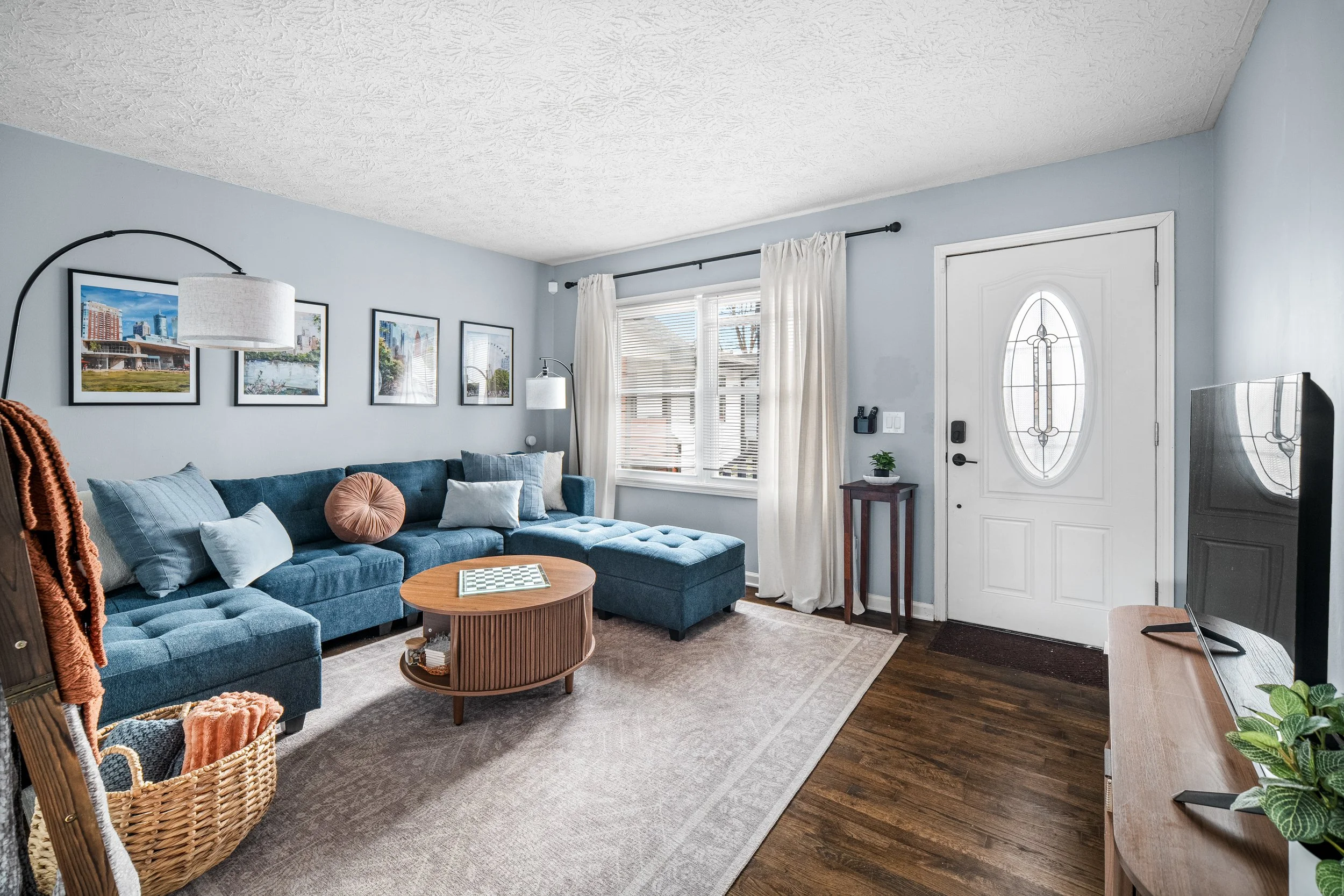 Living room with blue sectional sofa, throw pillows, coffee table, wall art, window with white curtains, TV, wooden floor, front door with oval window.
