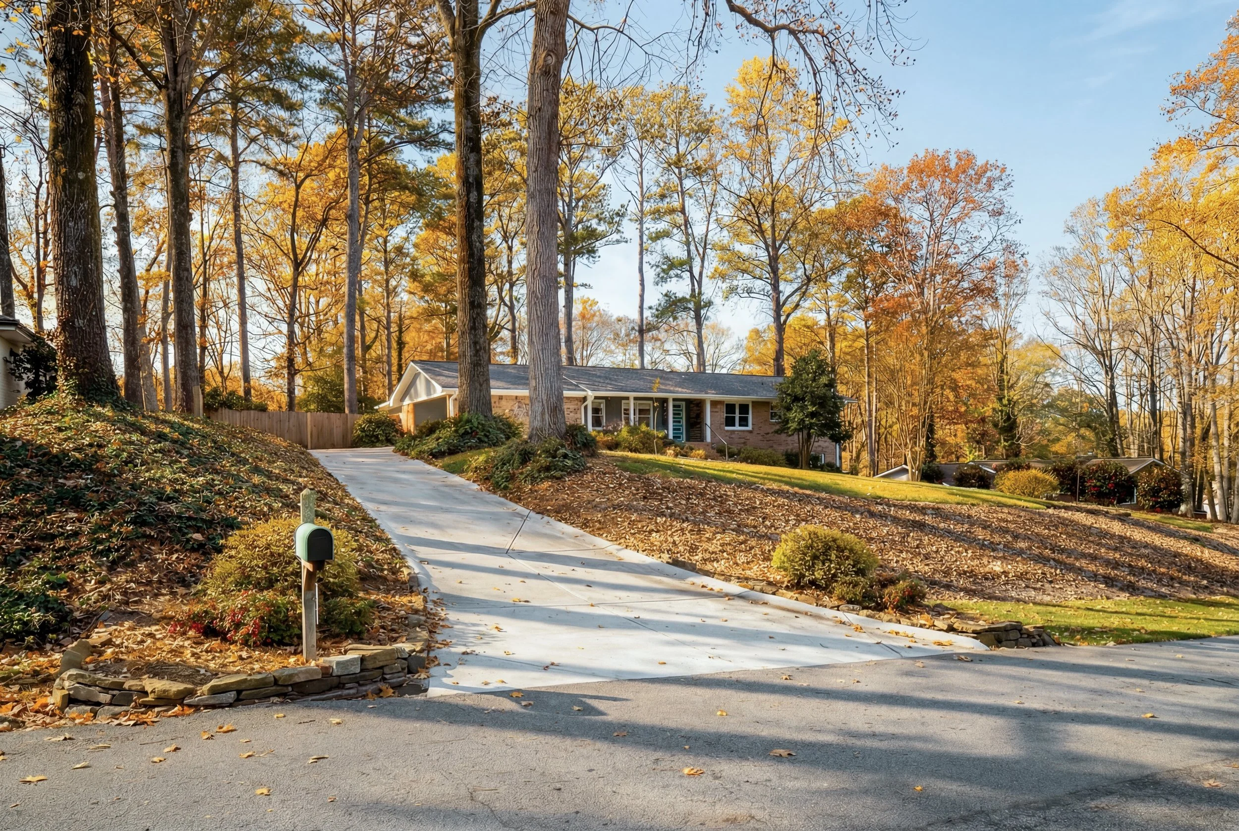 A suburban house on a hill during fall, with a concrete driveway, surrounded by trees with orange and yellow leaves, and a front yard with bushes.
