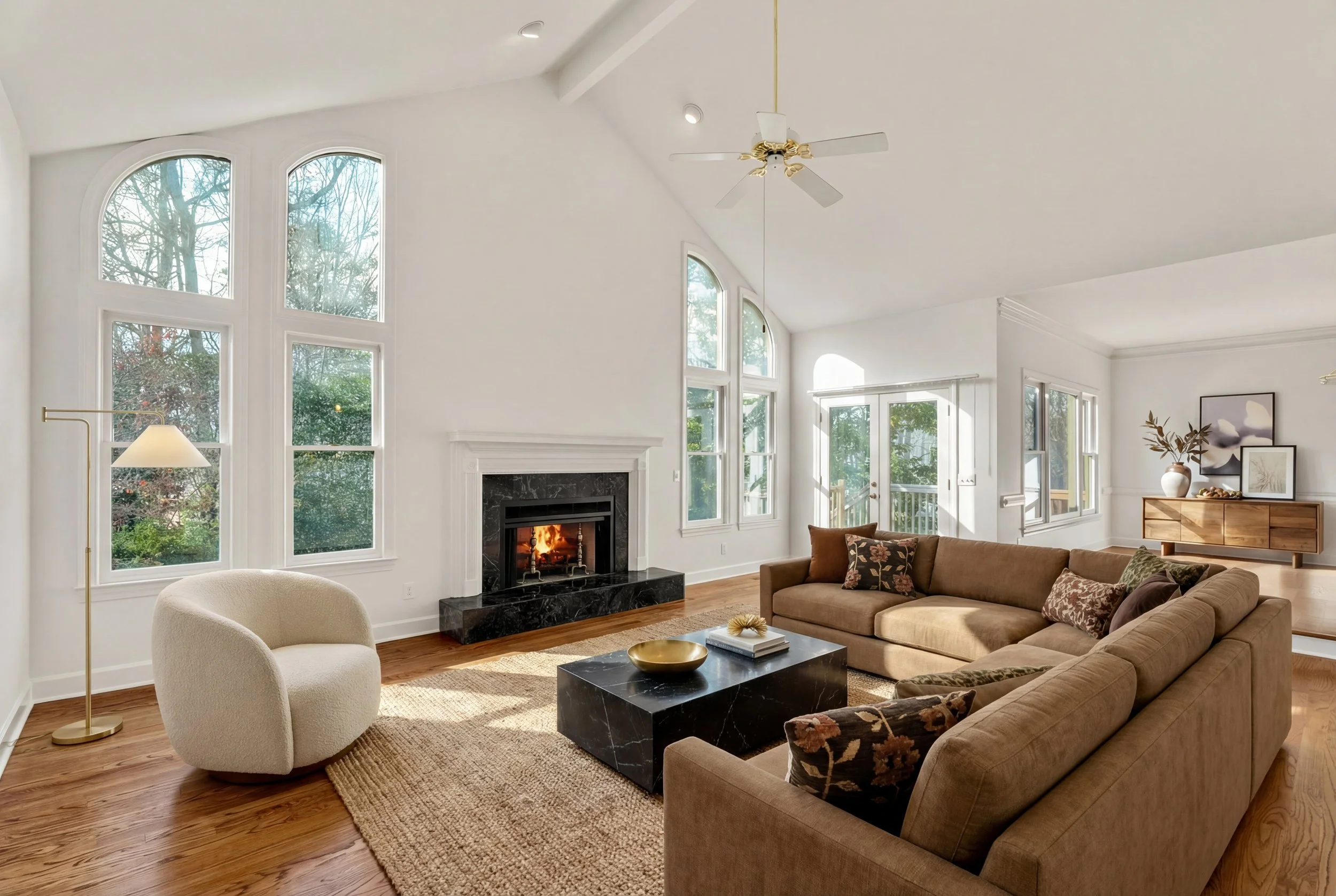 Living room with large arched windows, fireplace, beige sectional sofa with pillows, white armchair, black marble coffee table, wooden sideboard with decor, hardwood floor, and ceiling fan