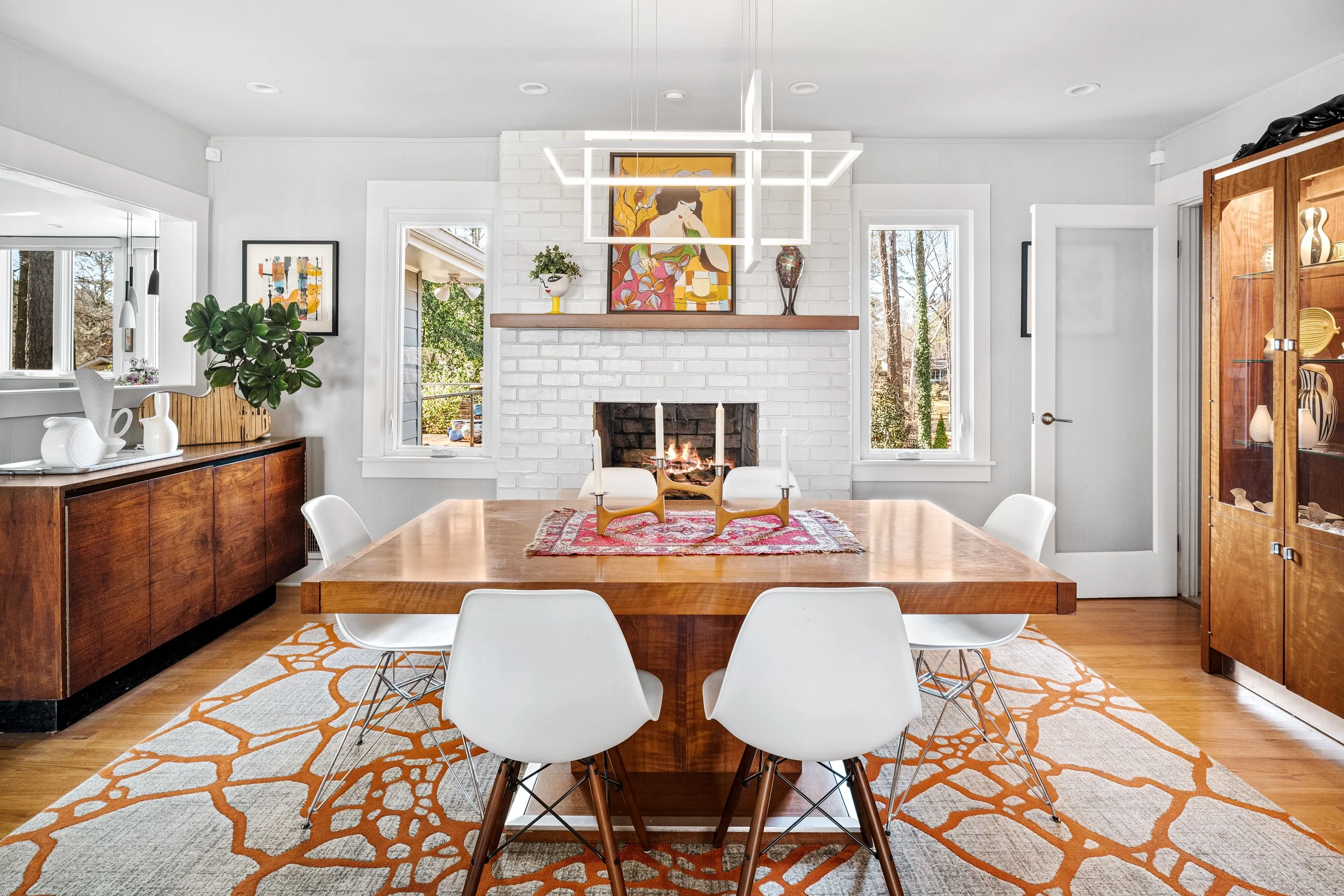 A dining room with a wooden table and six white chairs, a white brick fireplace, decorative artwork above the mantel, a patterned rug, wooden sideboards, and large windows with a view of trees outside.