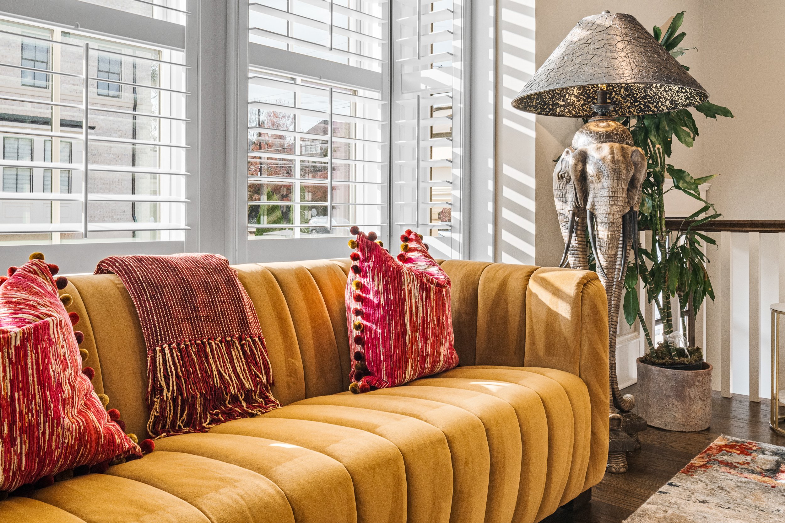 Interior of a living room featuring a mustard yellow upholstered sofa with three red patterned pillows with pom-poms, a large window with white shutters, and a decorative lamp with an elephant-shaped base and a metallic shade, next to a green potted 