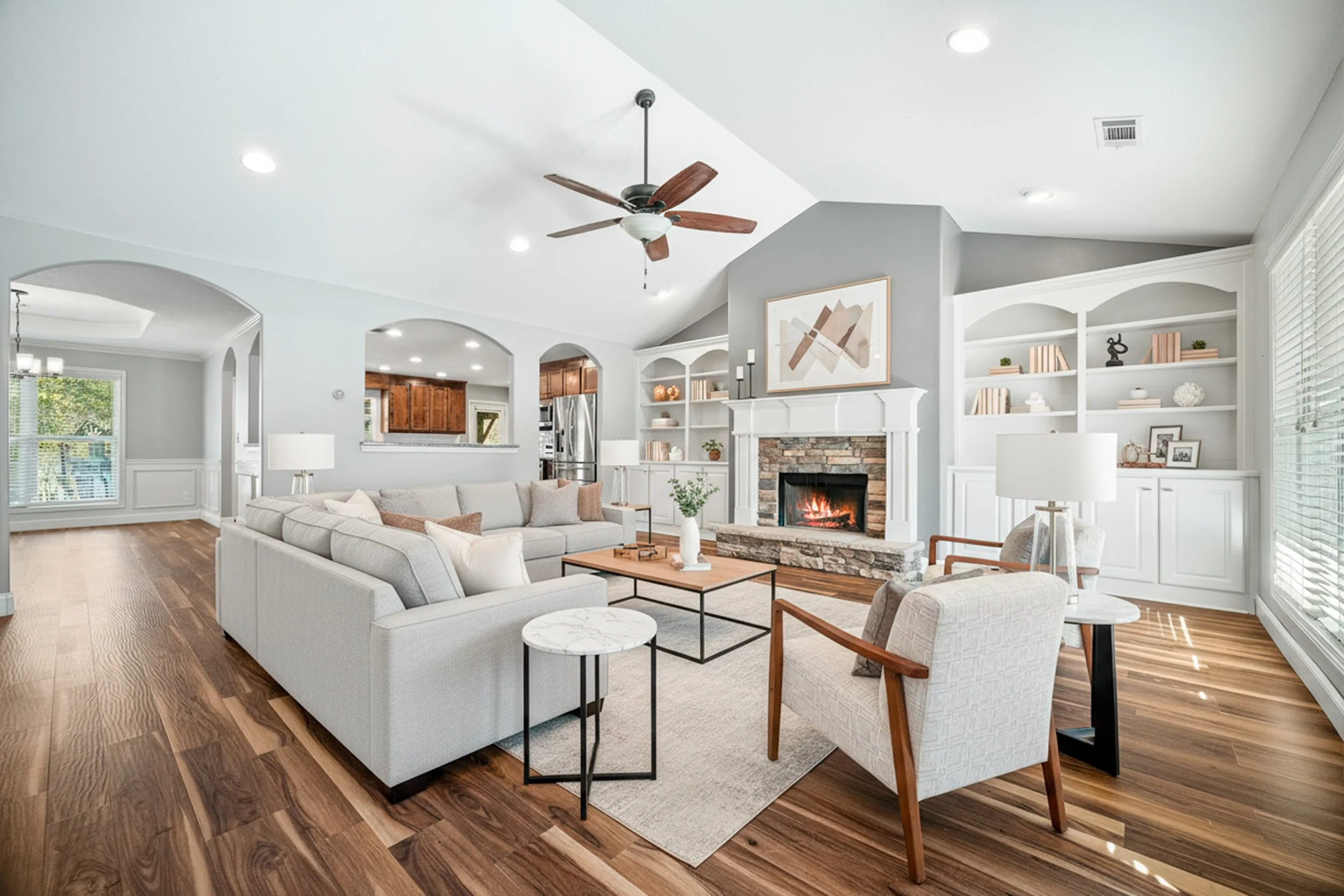 Living room with a white sofa, armchair, wooden coffee table, fireplace, built-in bookshelves, hardwood floors, ceiling fan, and large windows.