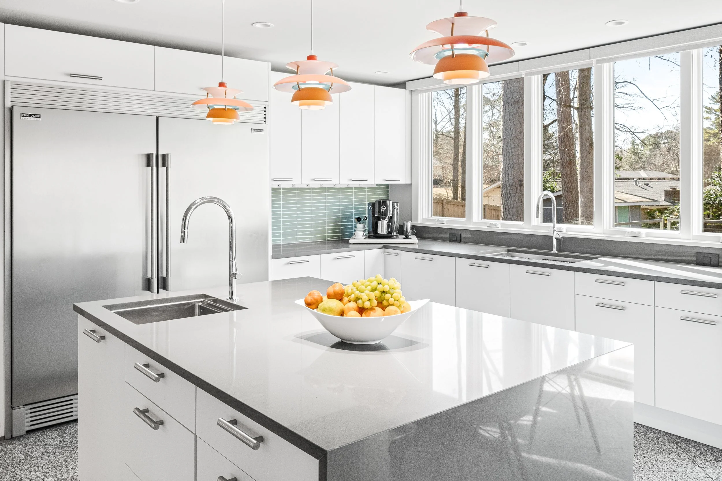 Modern white kitchen with large windows, white cabinets, gray countertops, and a central island with a bowl of fresh fruit.