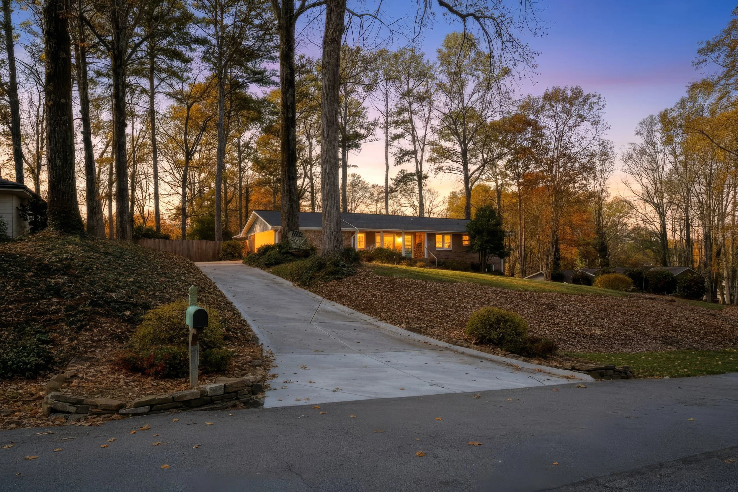 A house on a hill at sunset with large trees surrounding it, a concrete driveway leading up to the house, and a mailbox in the foreground.
