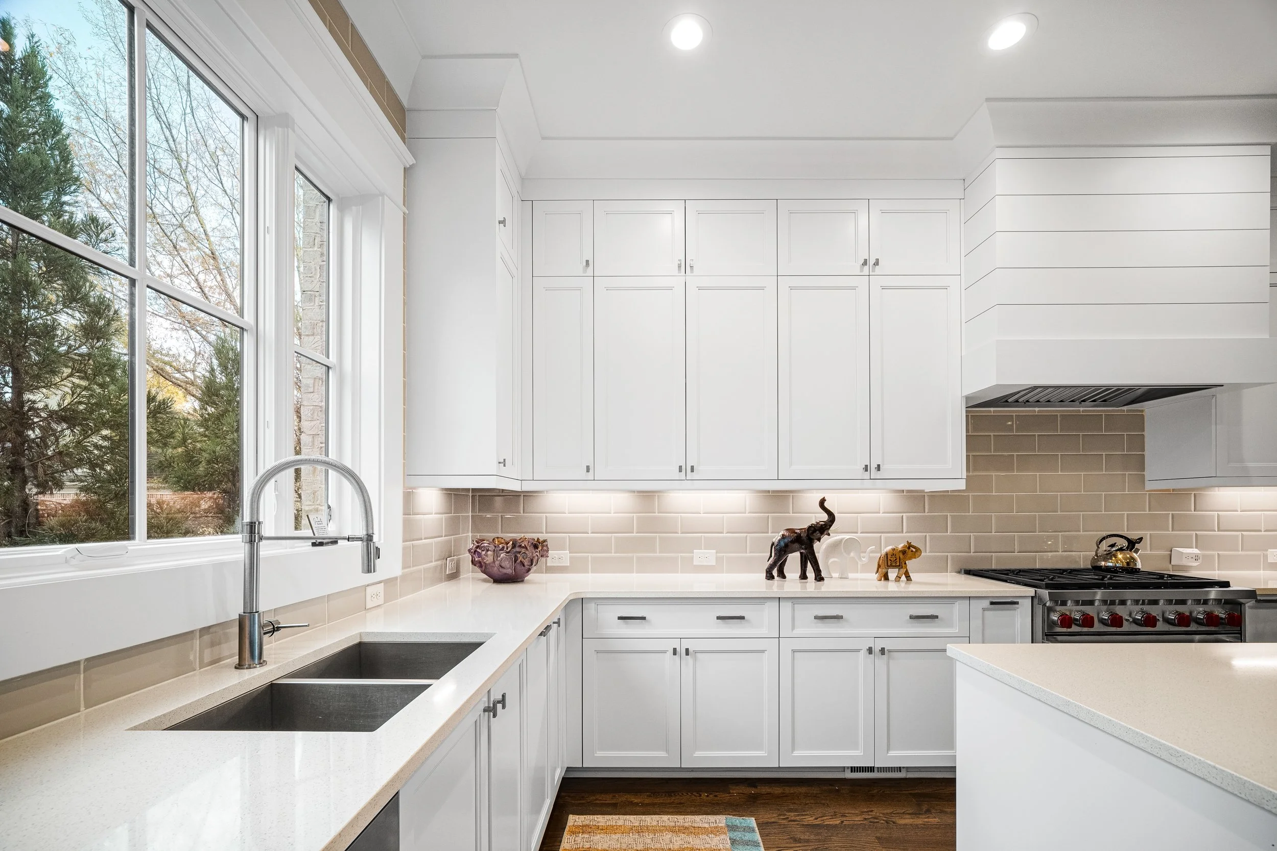 Kitchen with white cabinets, beige subway tile backsplash, double sink, and a stove with a kettle, decorated with small elephant figurines on the counter, and large windows showing trees outside.