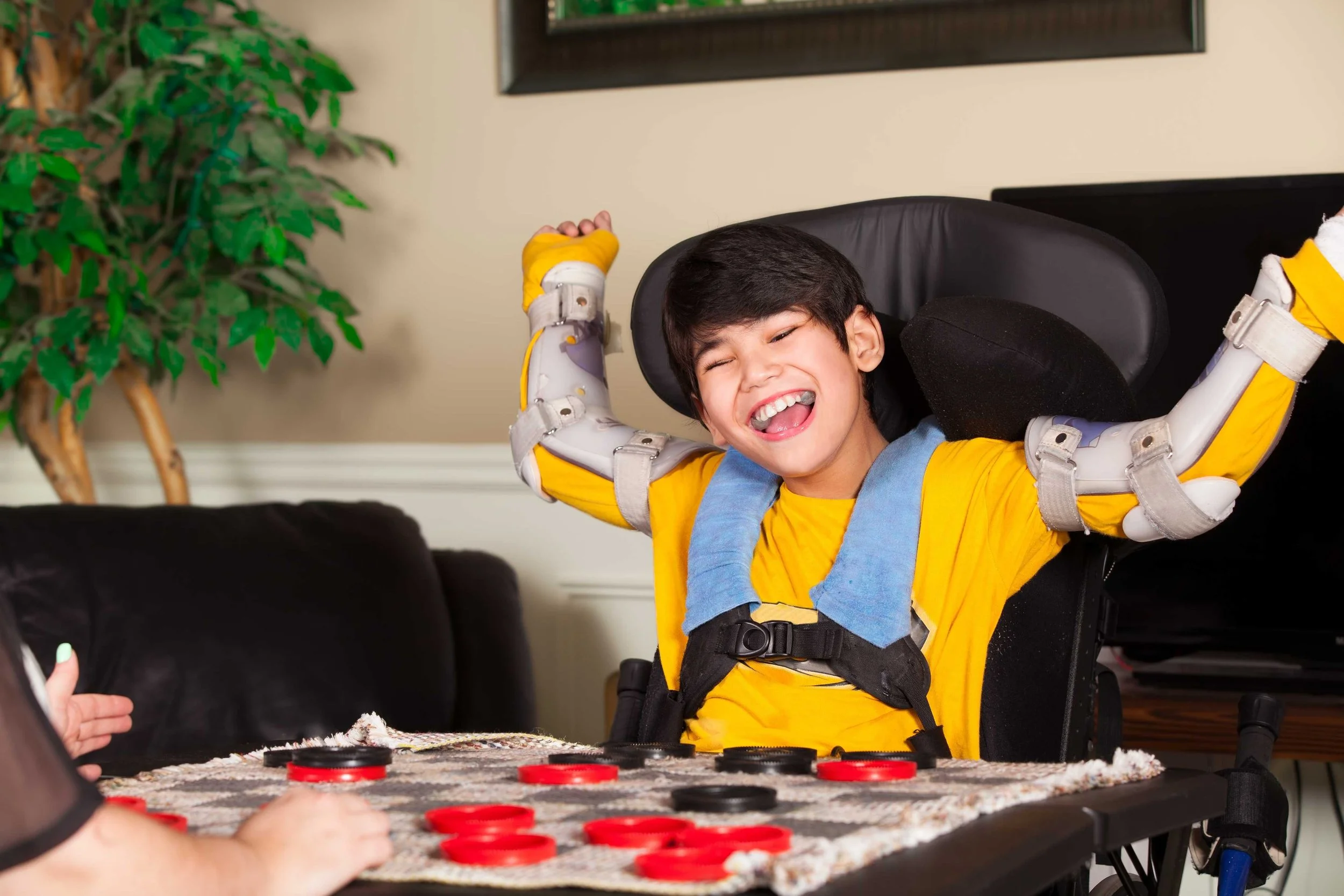 Smiling child in a wheelchair raising their arms in excitement while playing a game of checkers indoors.