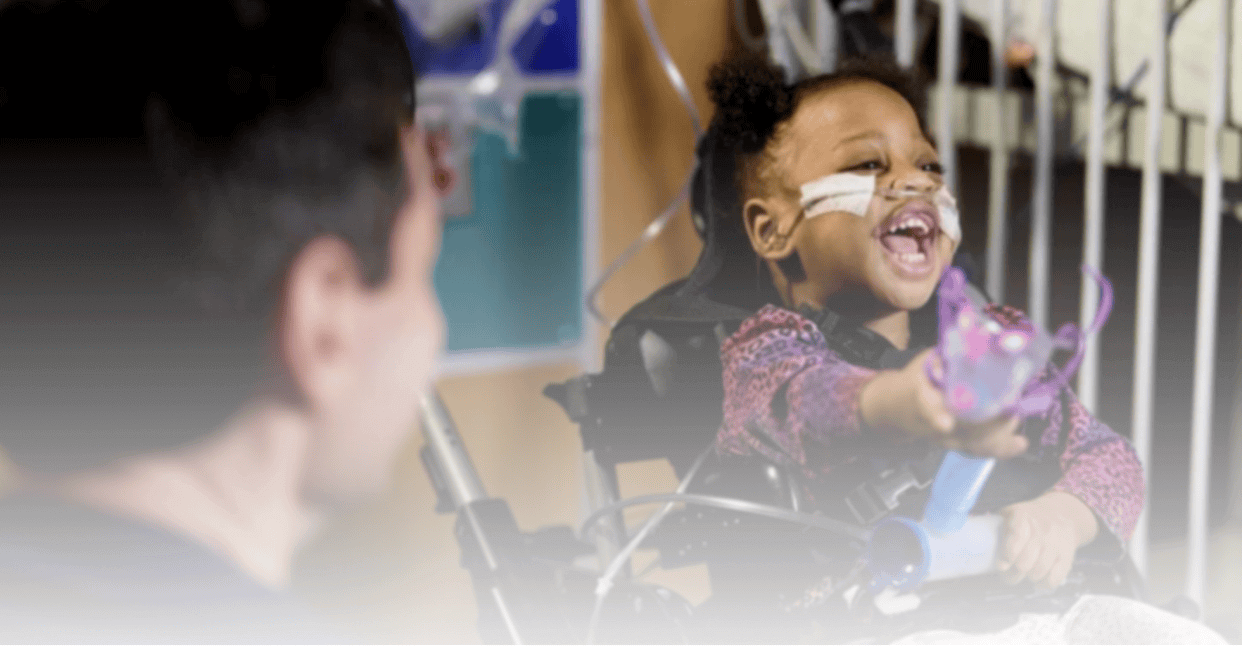 Young girl in a wheelchair laughing and reaching out while interacting with a caregiver in a medical setting.