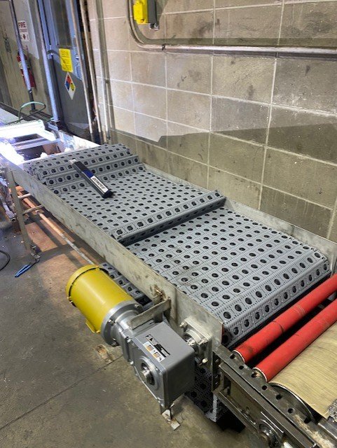 A conveyor belt system in an industrial setting, featuring a gray plastic modular belt and motor, positioned against a concrete block wall. The belt has black circular patterns and adjacent red rollers.