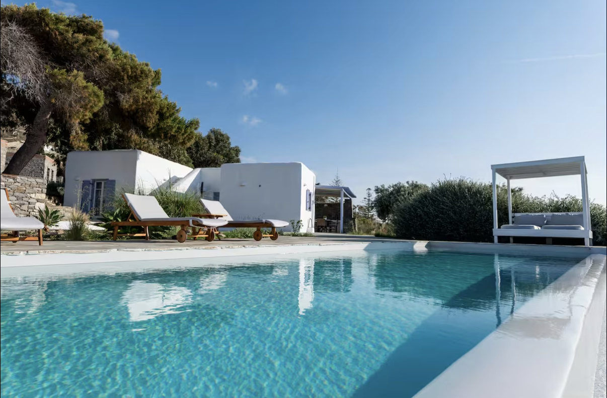 Scenic view of a luxurious outdoor swimming pool with white lounge chairs, cabana, and lush greenery under a clear blue sky.