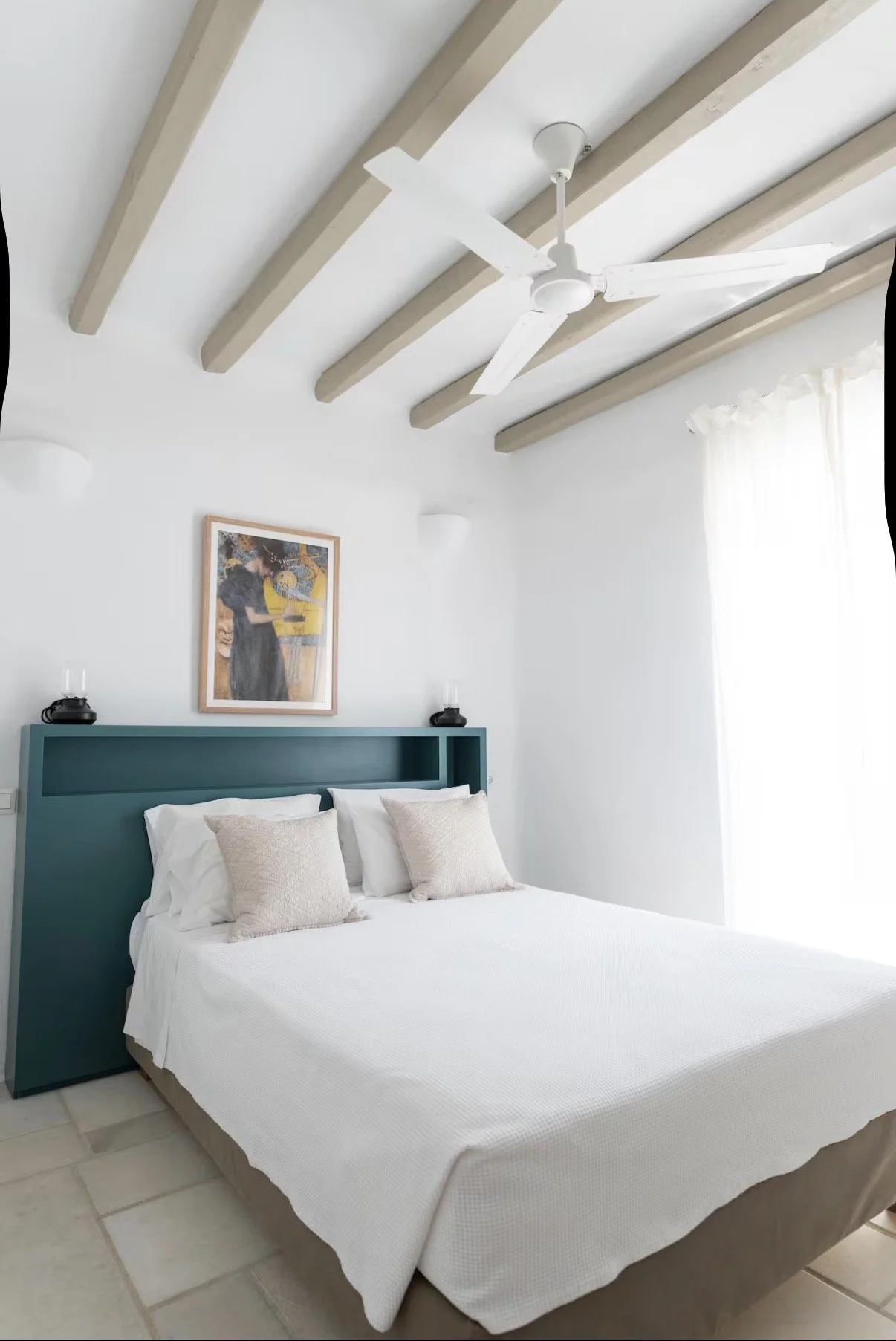 A bedroom with a white bed, beige pillows, a teal headboard, a painting above the bed, and a ceiling fan with white blades. There are two wall-mounted lights, and a window with white curtains.