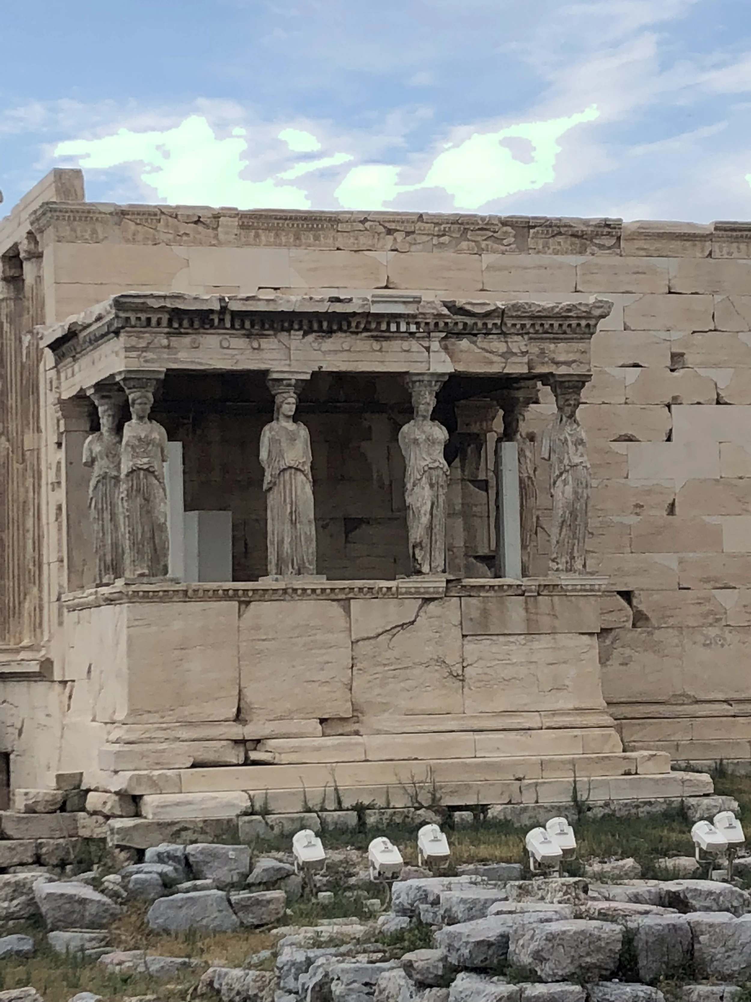 Ancient Greek temple with marble columns and architecture, partly damaged, with modern white security lights at the base, set against a cloudy sky.
