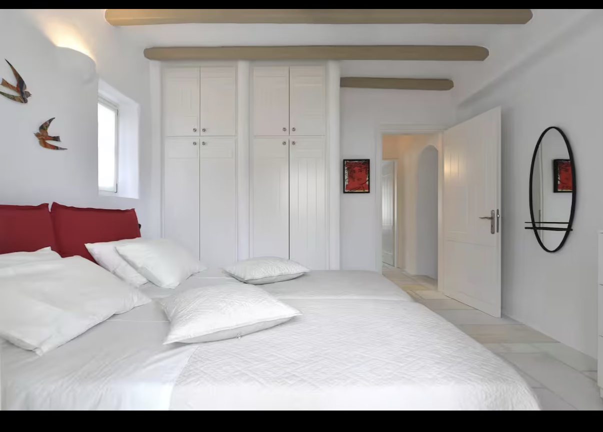 A modern, minimalistic bedroom with a white bed, red headboard, white pillows, wall art of flying birds, a small window, white closet with closed doors, a doorway leading to another room, a round mirror on the wall, and wooden ceiling beams.