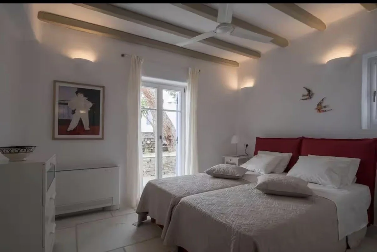 A cozy bedroom with two beds featuring red headboards, white bedding, and pillows. There's a white nightstand with a small lamp, a framed artwork on the wall, and decorative birds on another wall. The room has beam accents on the ceiling, soft lighting, and a glass door leading outside with white curtains.