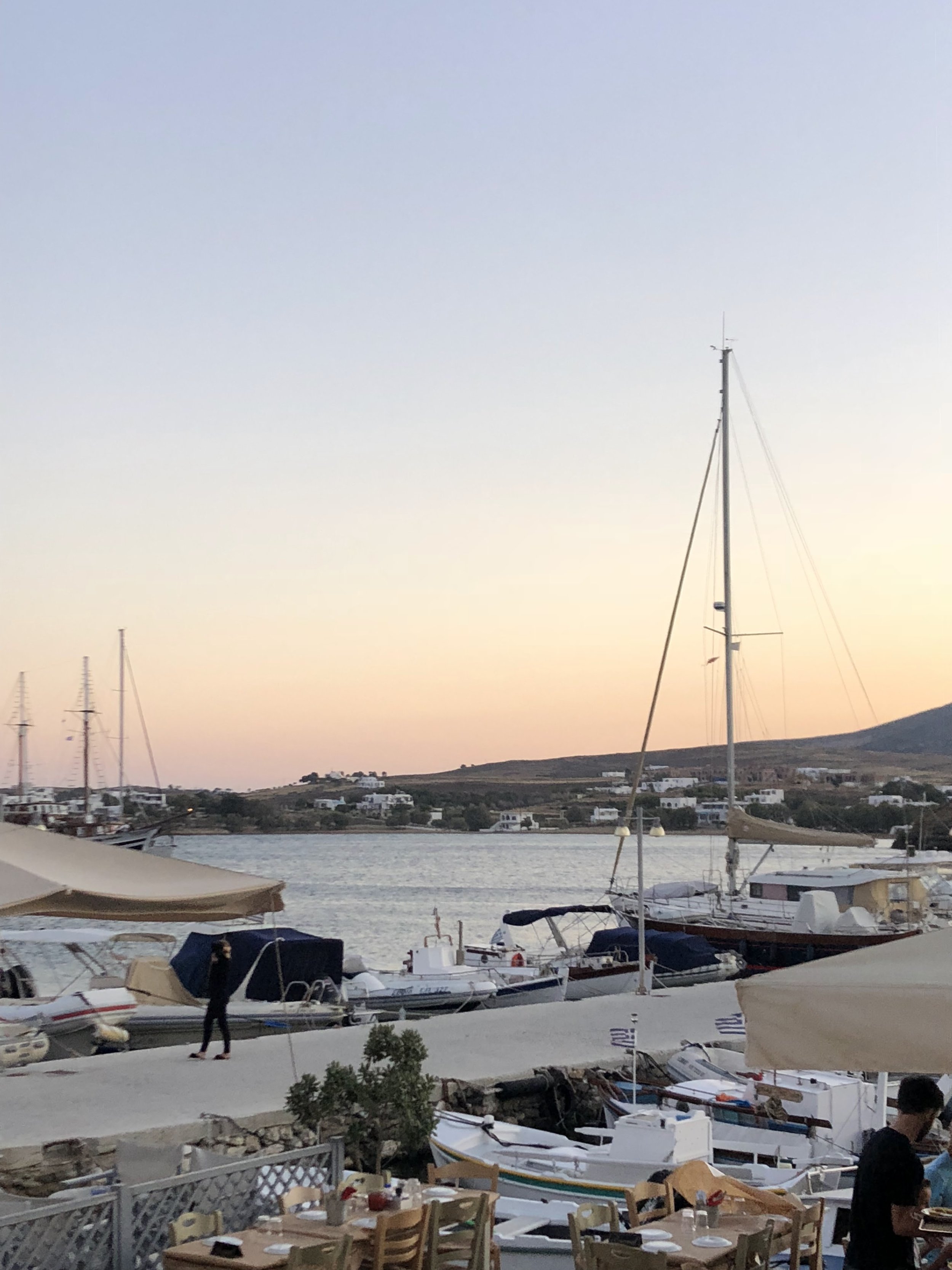 A marina at sunset with several sailboats docked, including a prominent sailboat with a tall mast. In the foreground, people are walking and dining outdoors at a restaurant with tables and chairs, and some tents or umbrellas are visible. The background features a hillside with white buildings and a fading sky with pastel colors.