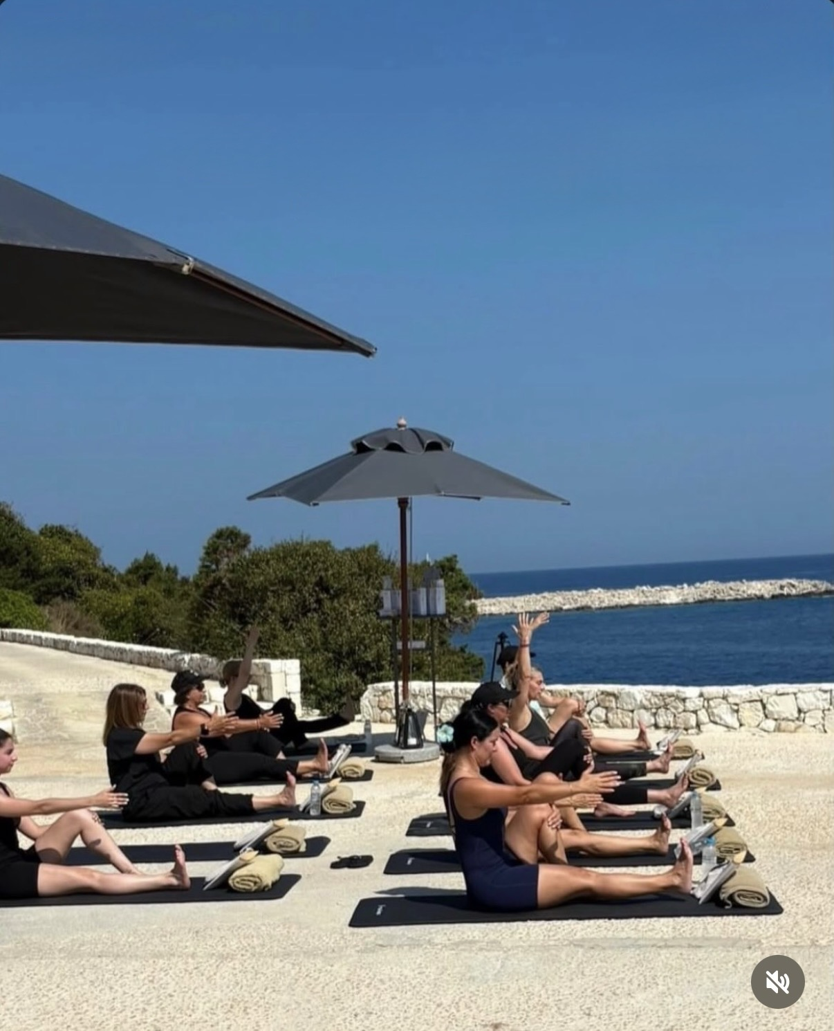 People participating in an outdoor yoga class on a beachside patio, sitting on yoga mats with their arms extended towards the sky, overlooking the ocean with some umbrellas providing shade.