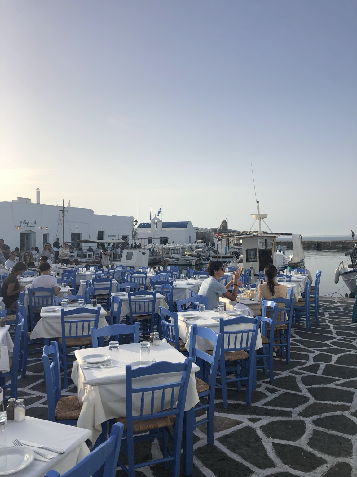 Open-air seaside restaurant with white tablecloths and blue chairs, overlooking boats docked at the harbor with white buildings and a stone pier, during sunset.