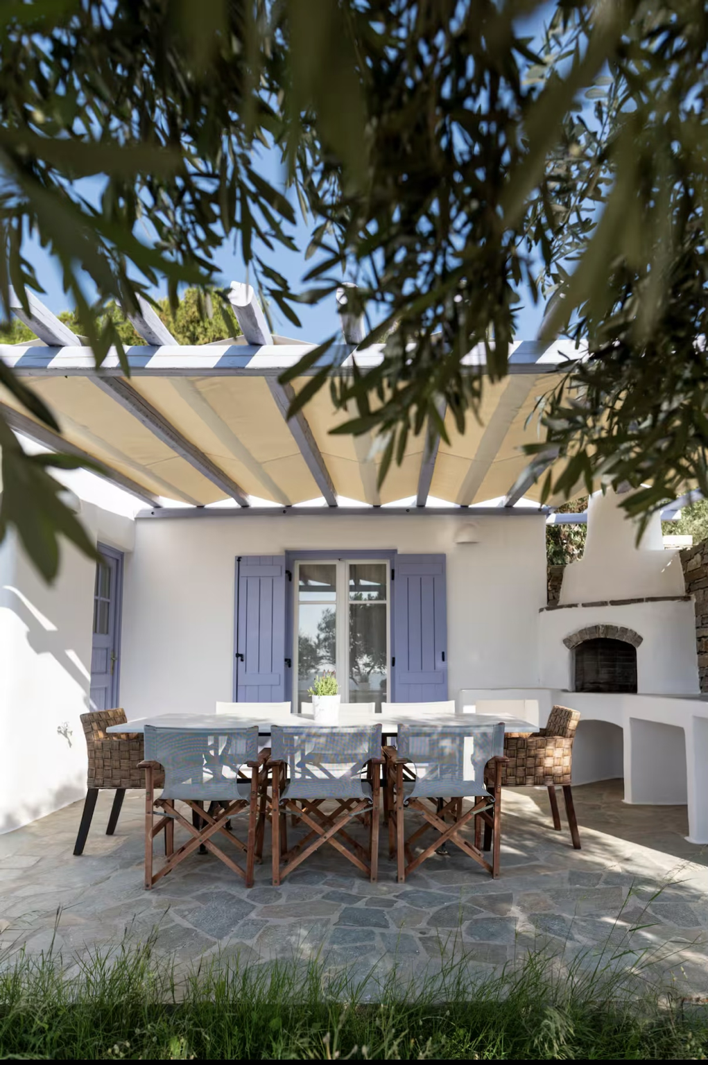 Exterior patio of a white house with blue shutters, a stone floor, a dining table with seven chairs, a small plant on the table, and a built-in outdoor oven, partially shaded by a pergola and trees.
