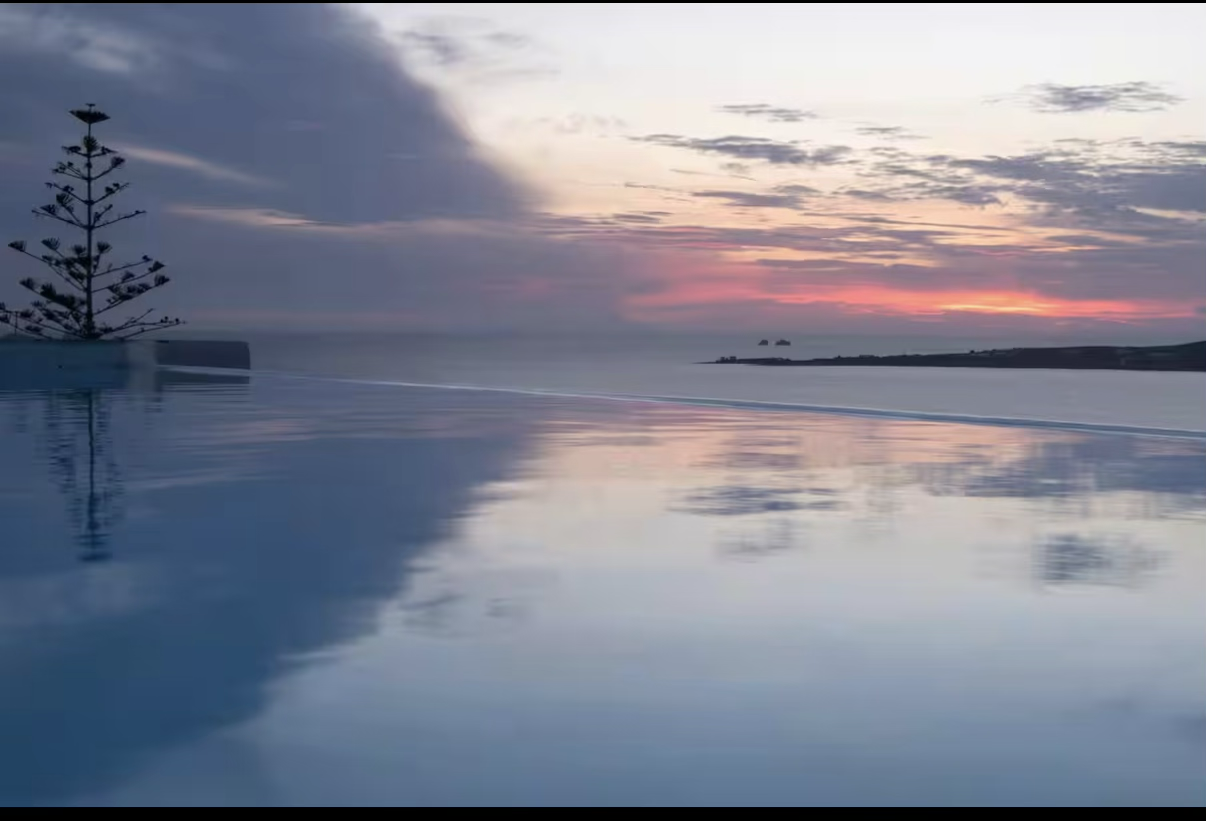 A sunset over the ocean with a partially cloudy sky. A tree is reflected in a calm, mirror-like pool of water in the foreground.