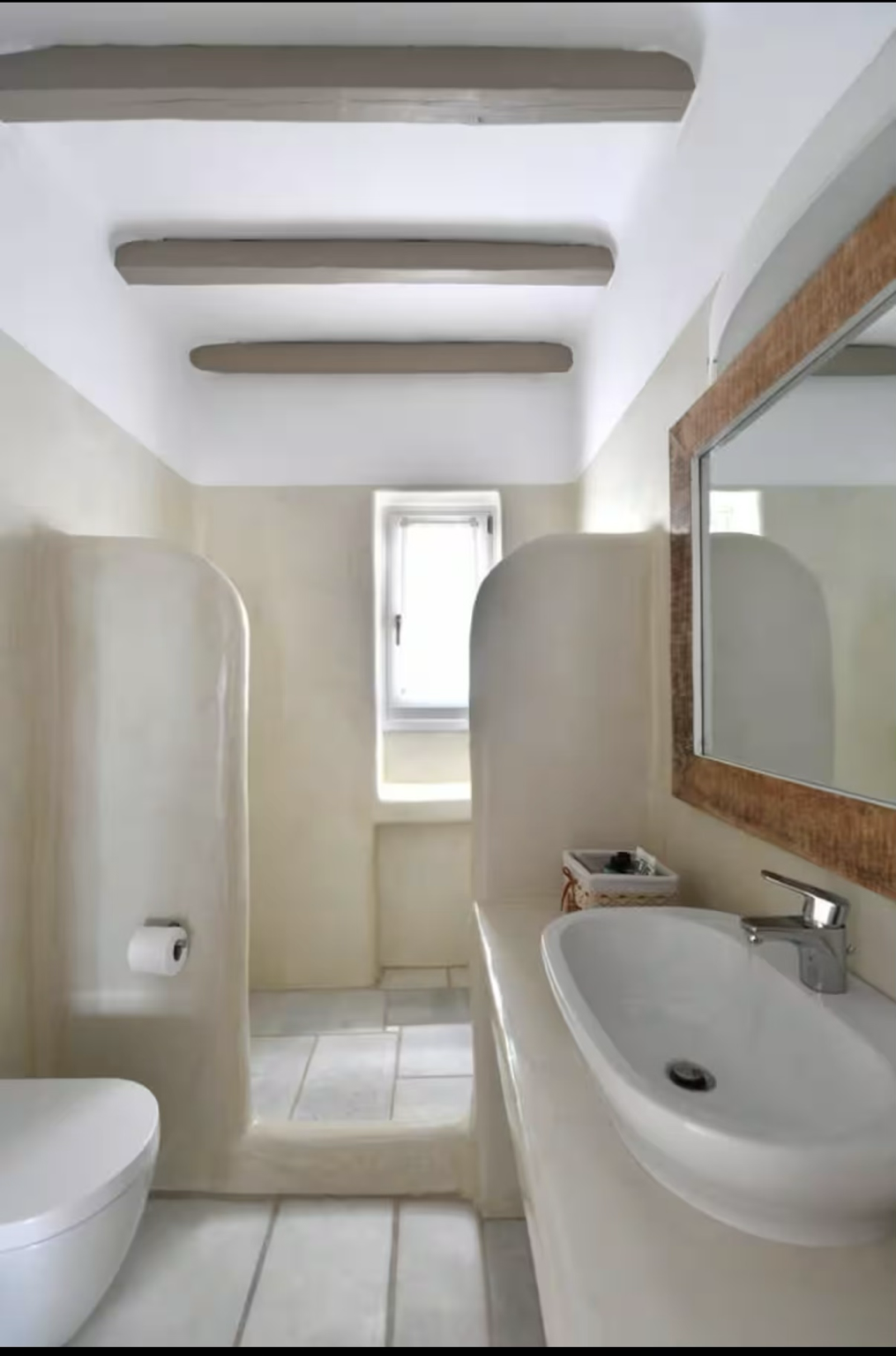 A small bathroom with a toilet, a sink with a mirror, and a small window. The bathroom has light-colored walls, tiled flooring, and a ceiling with wooden beams.