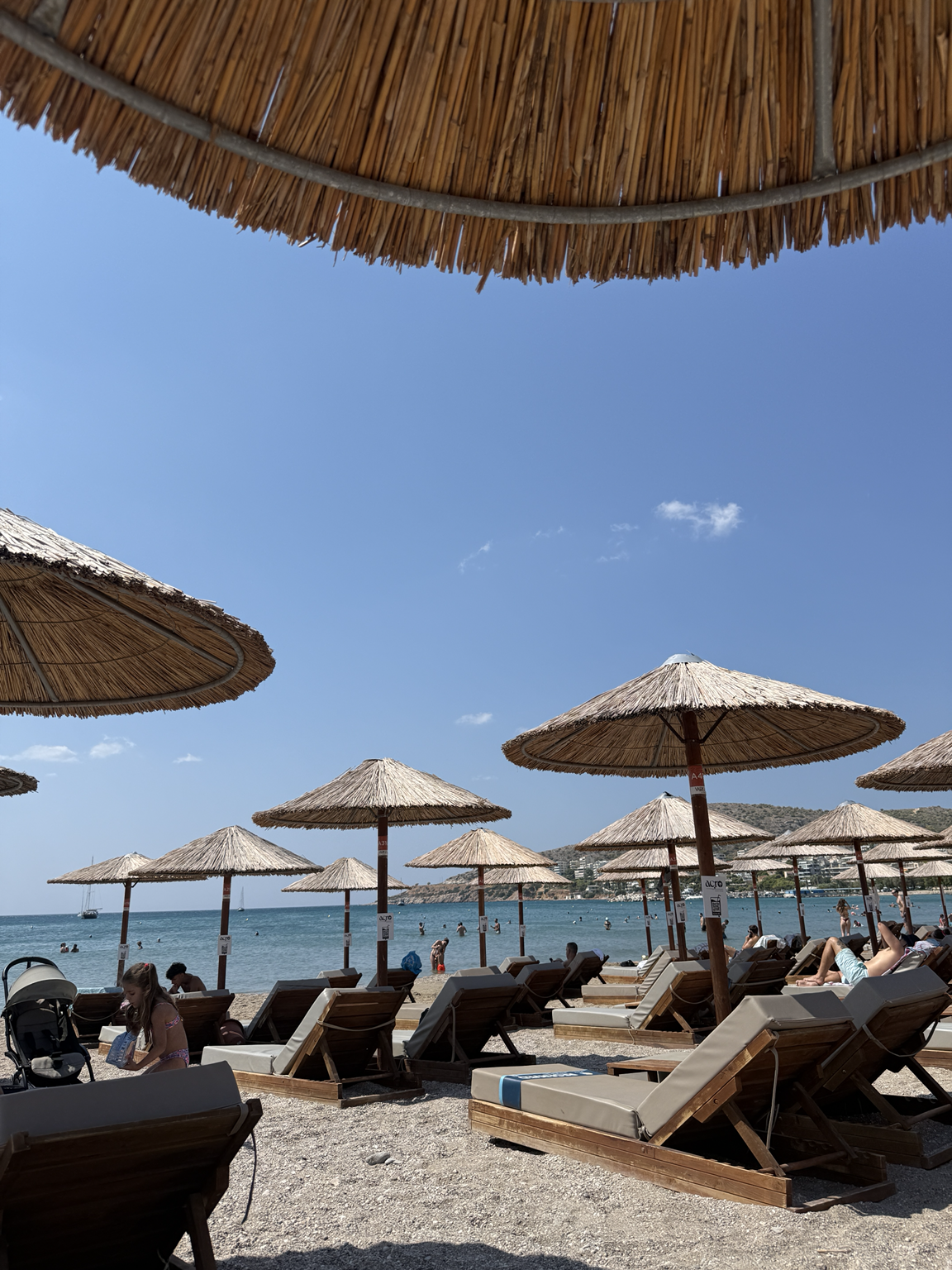 Beach with thatched umbrellas, chairs, and people swimming in the ocean under a clear blue sky.