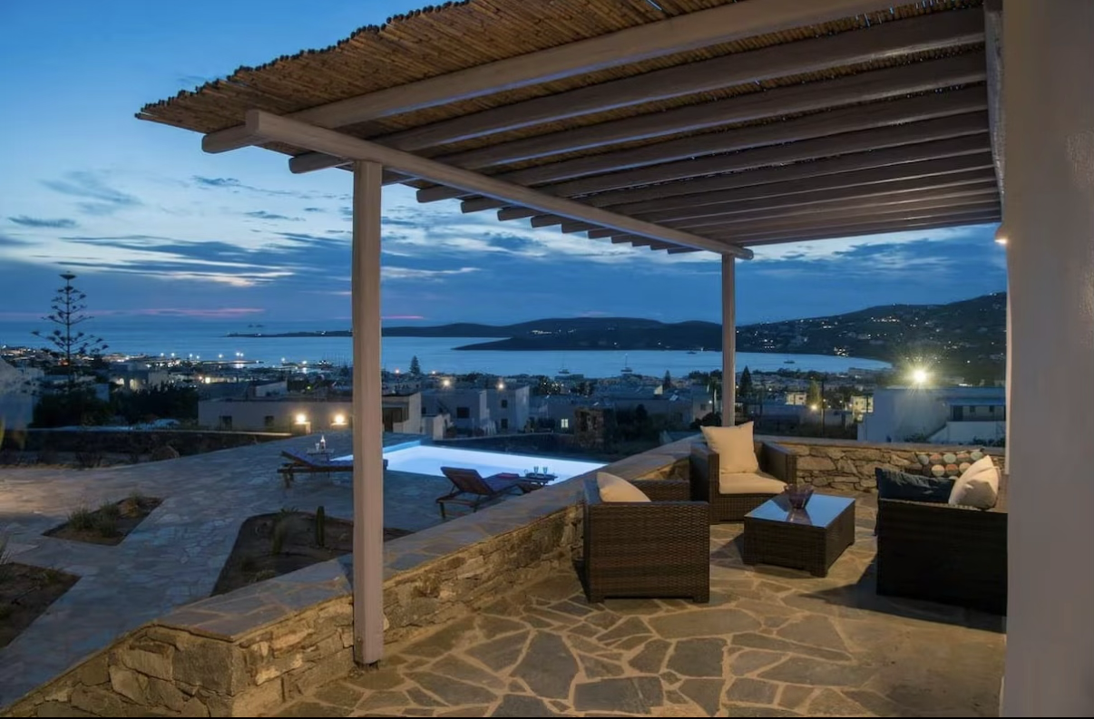 A cozy outdoor seating area on a stone patio at dusk, with a view of a coastal town, distant mountains, and a pool illuminated nearby.