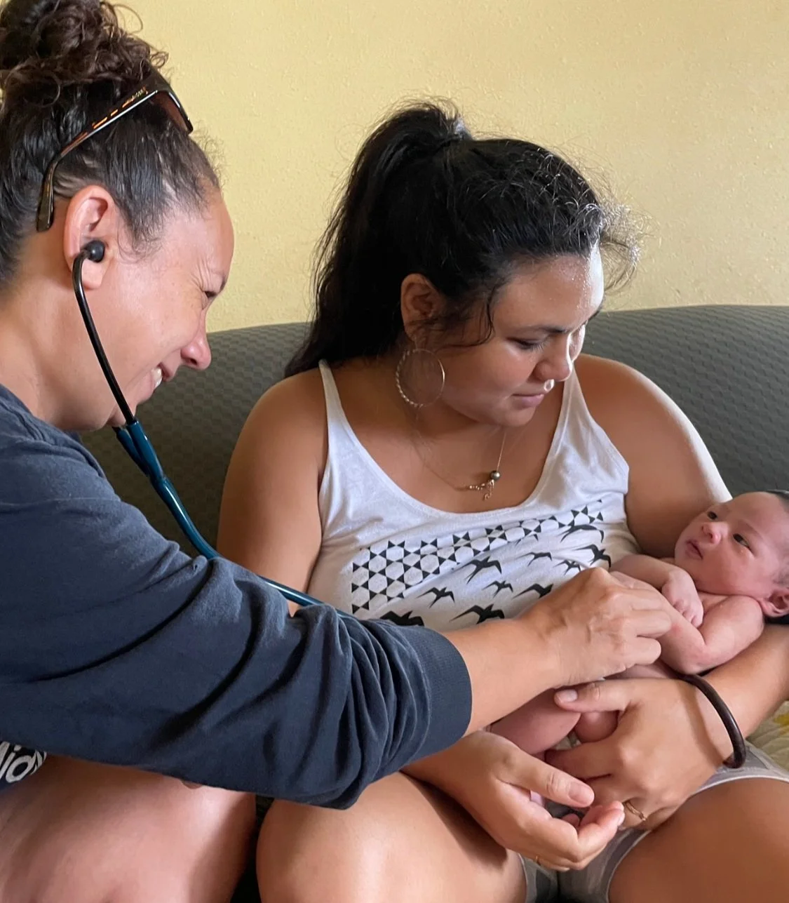 A nurse using a stethoscope to examine a newborn held by a woman sitting on a couch.