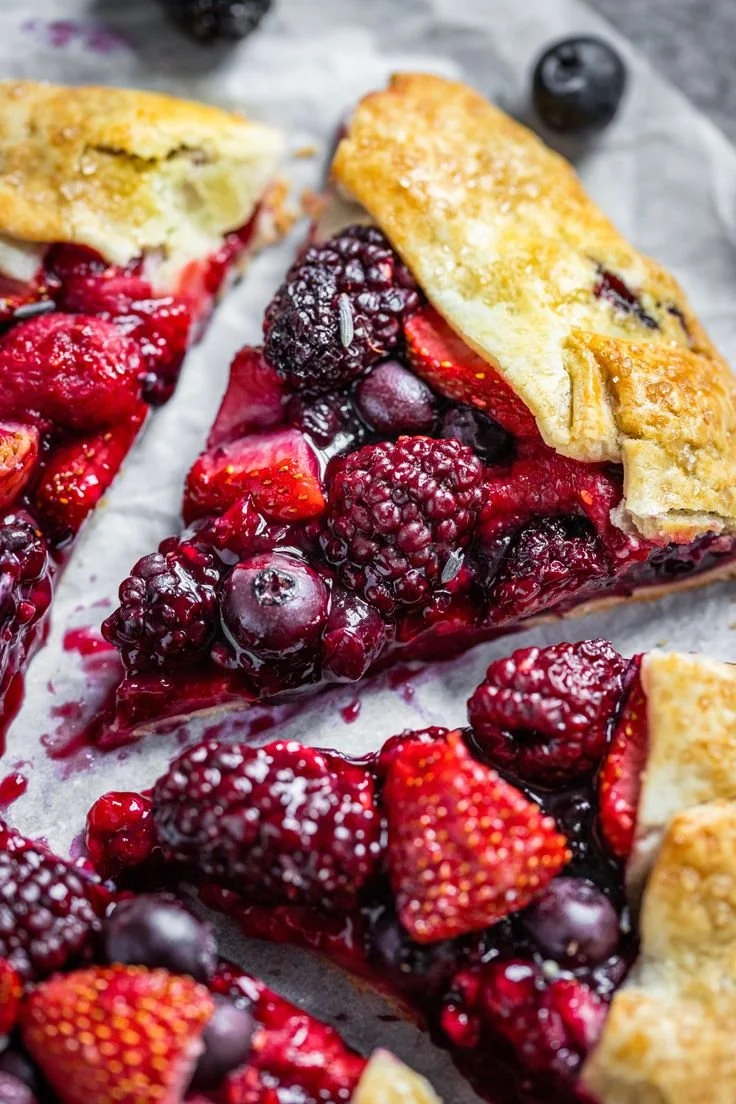 Close-up of a berry pie with a golden crust, filled with mixed blackberries, strawberries, and raspberries, some spilled on parchment paper.