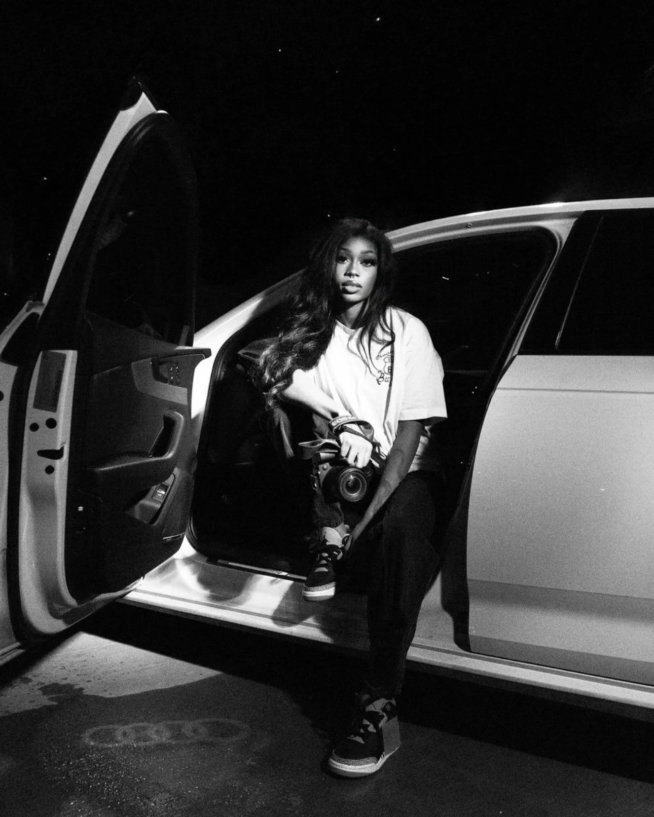 Black and white photo of a woman sitting in the open door of a car at night, holding a camera, wearing casual attire.